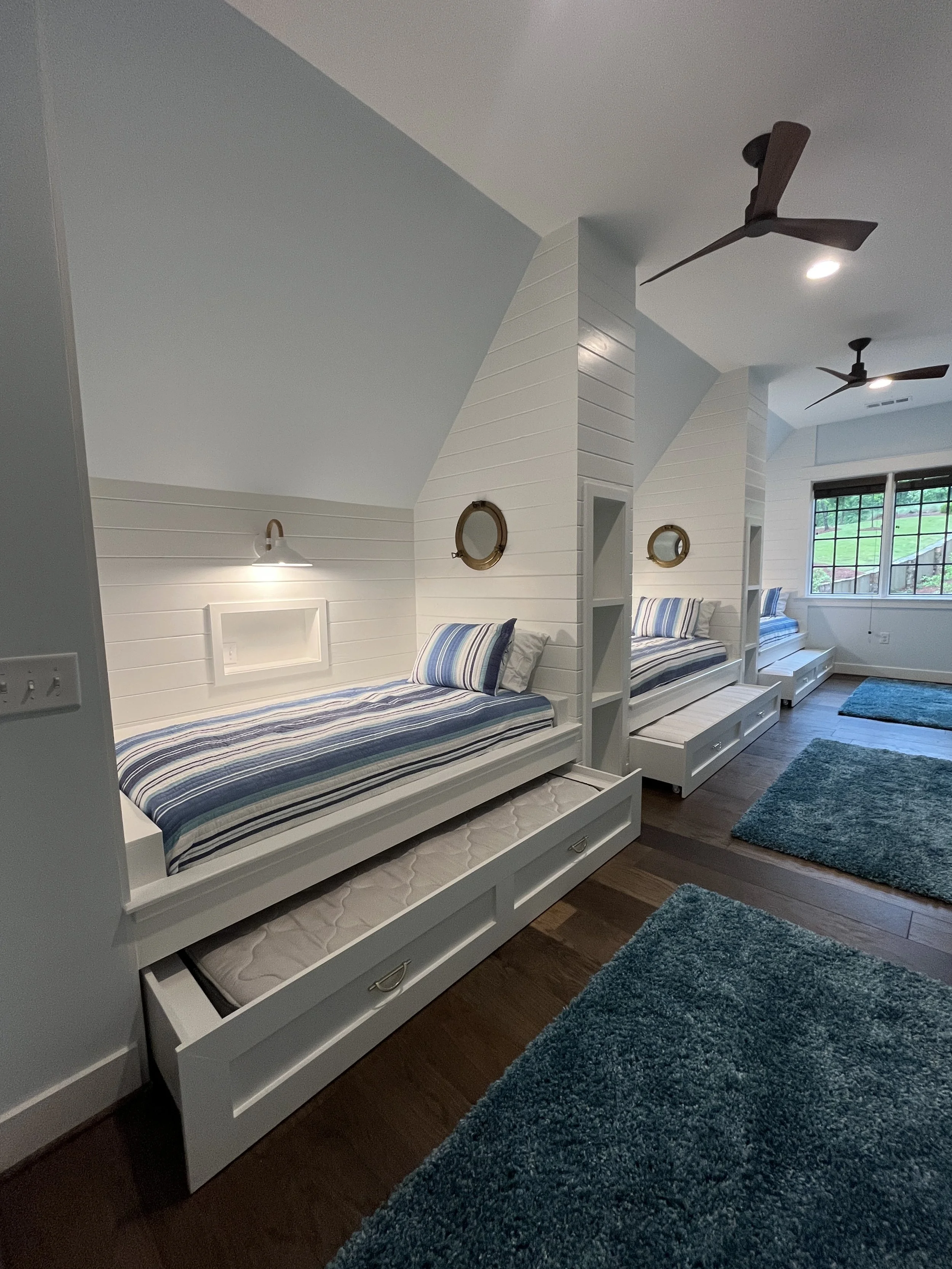 A bedroom with three built-in beds with striped bedding, under sloped ceilings, featuring white wooden panel walls, blue area rugs, and ceiling fans.