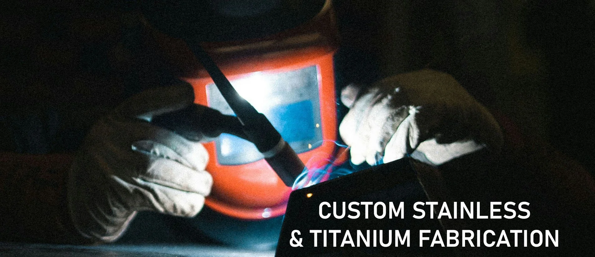 Hands wearing gloves holding a welding torch and working on a stainless steel object in a workshop, with the text 'Custom Stainless & Titanium Fabrication' overlayed.