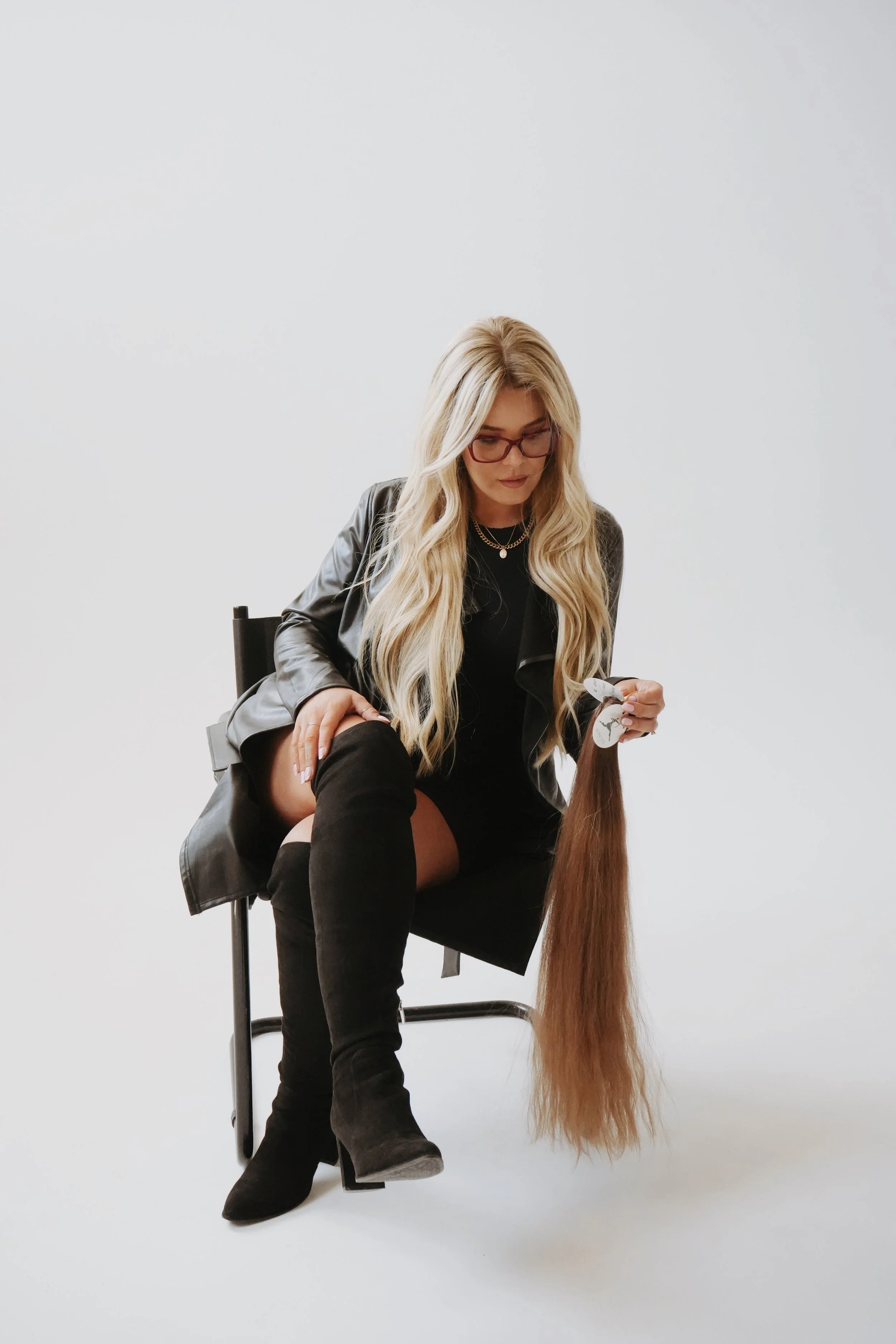 A blonde woman with long hair, wearing glasses, a black outfit, and a leather jacket, sits on a chair against a plain white background. She is holding a long strand of brown hair.