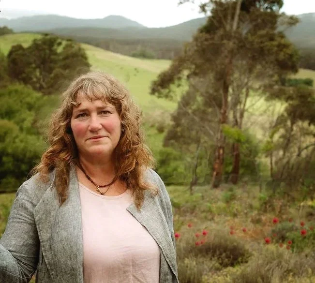 A woman standing outdoors in a green, hilly landscape with trees and red flowers, wearing a gray blazer and a light-colored shirt.