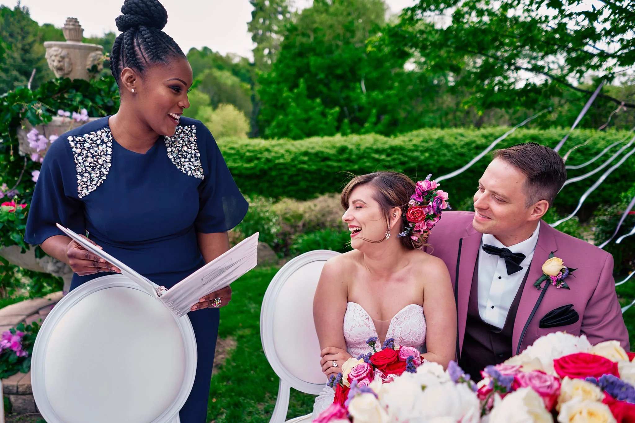 A woman with curly dark hair takes notes at a decorated banquet table with floral centerpieces, glasses, and place settings in a formal event space.