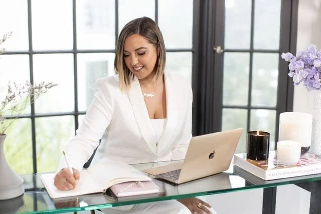 A woman in a white blazer is sitting at a glass desk in front of large windows, smiling while writing in a notebook next to her open laptop and decorative plants.