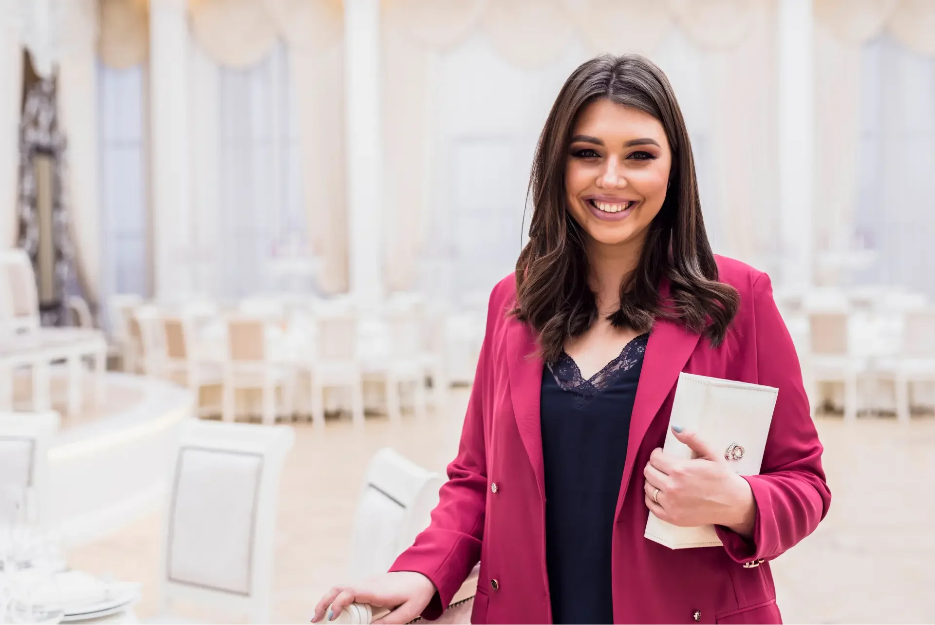 A woman with dark brown hair smiling, wearing a pink blazer over a navy top, holding a notebook, in a bright, elegant dining room with white chairs and large windows.