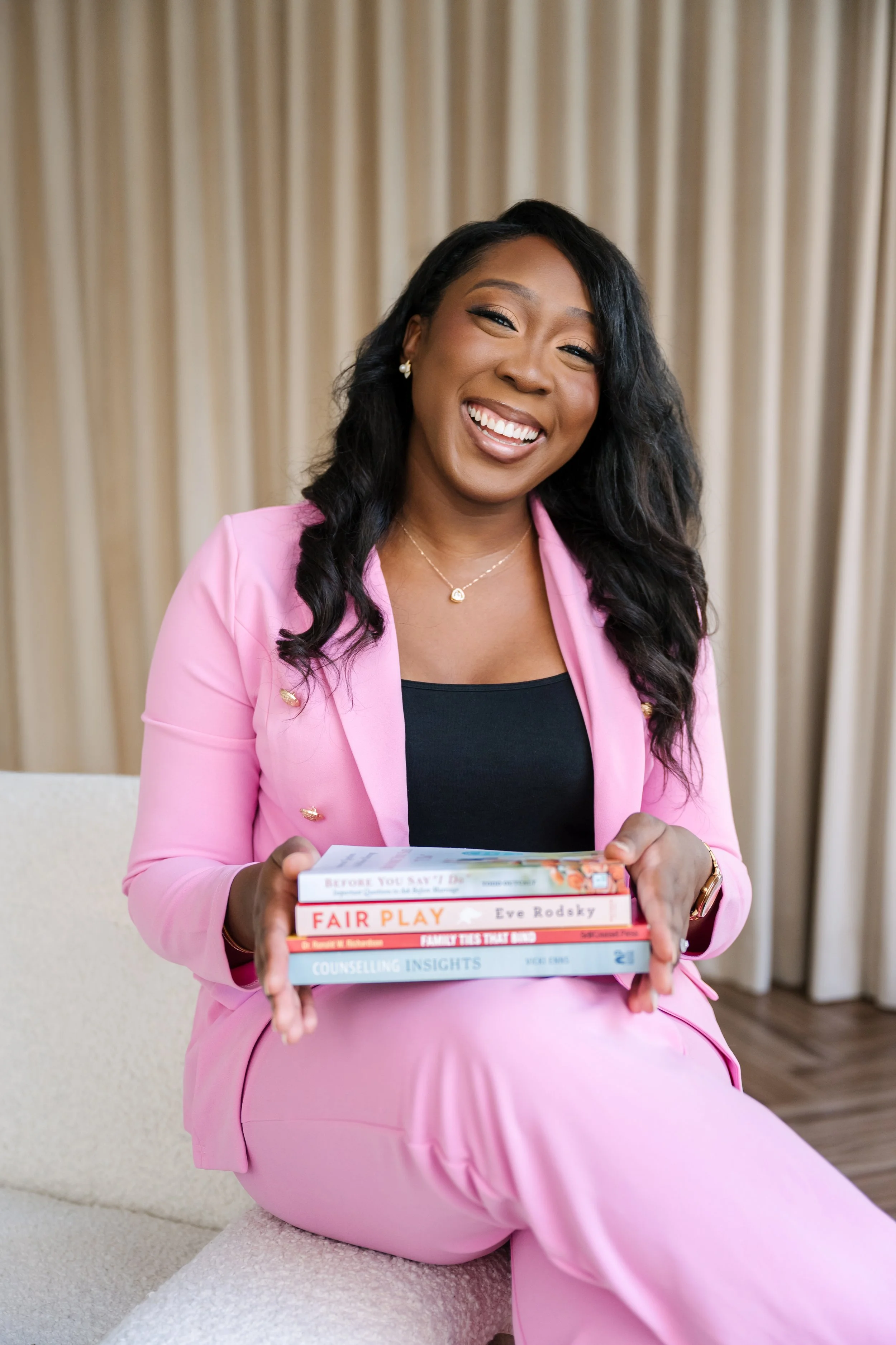 A smiling woman with dark curly hair, wearing a pink blazer and pants, sitting on a cream-colored sofa, holding a stack of five books on her lap, with beige curtains in the background.