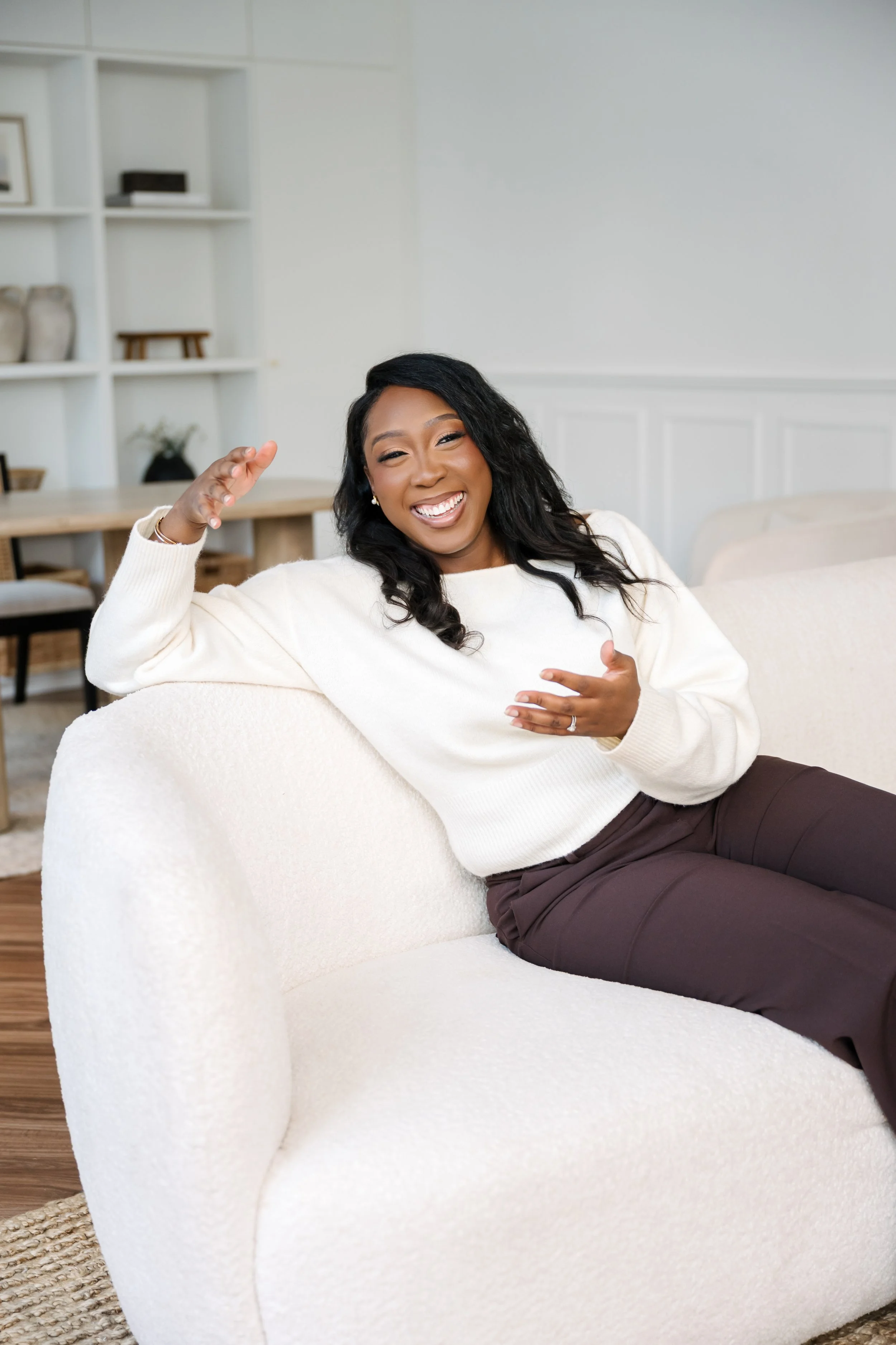 A woman with dark hair, smiling and laughing, sitting on a white sofa in a bright living room, wearing a white sweater and dark pants.