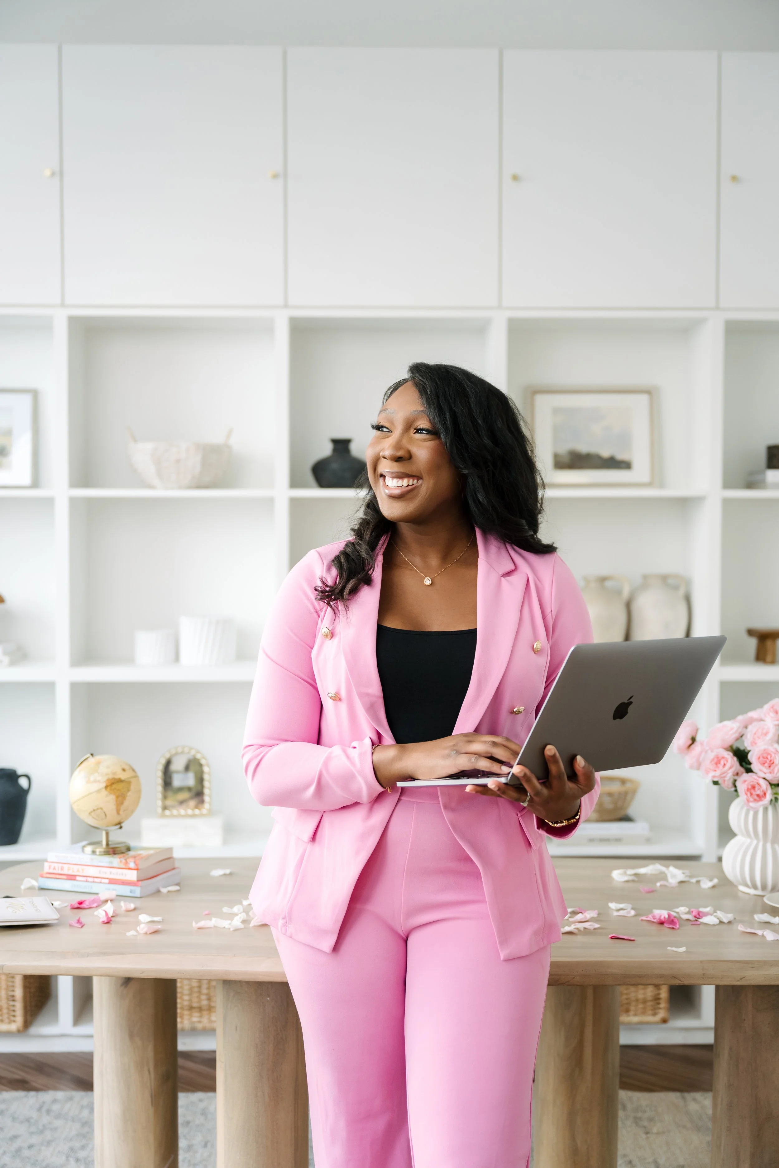 A woman in a pink suit holding a laptop, smiling, standing in a modern, bright room with white shelves and a decorated table.
