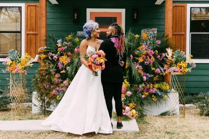 Two women at a wedding, one in a white wedding dress holding a bouquet, and the other in black clothes with colorful braids, smiling and looking at each other, standing in front of a green house with floral decorations.