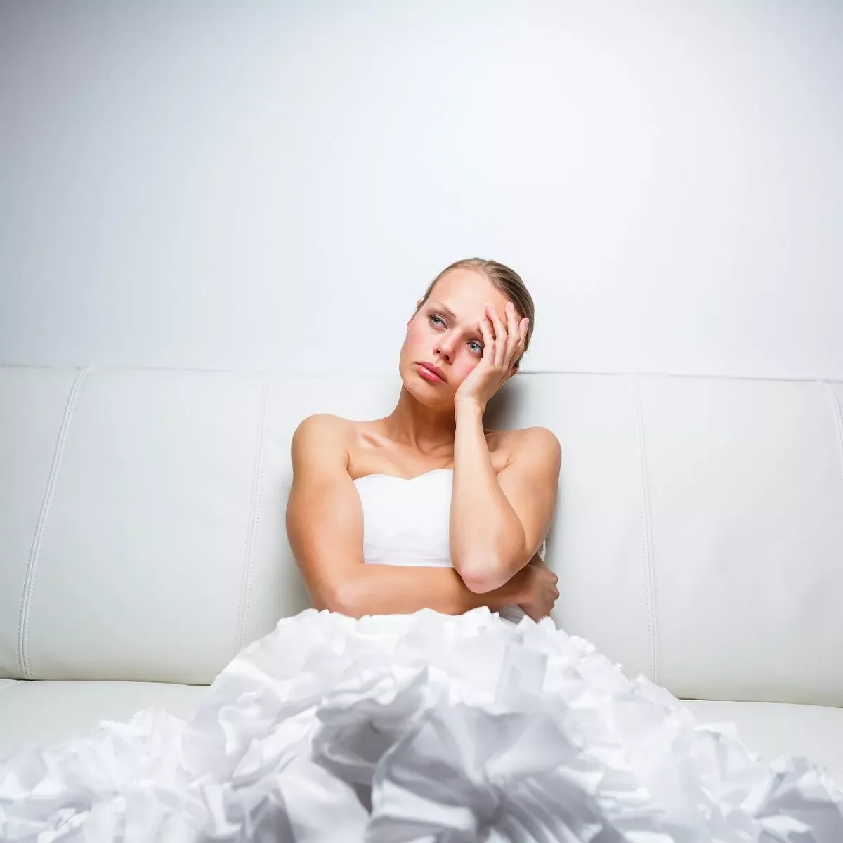A woman sitting on a white couch with a worried expression, holding her head with one hand, surrounded by crumpled papers.