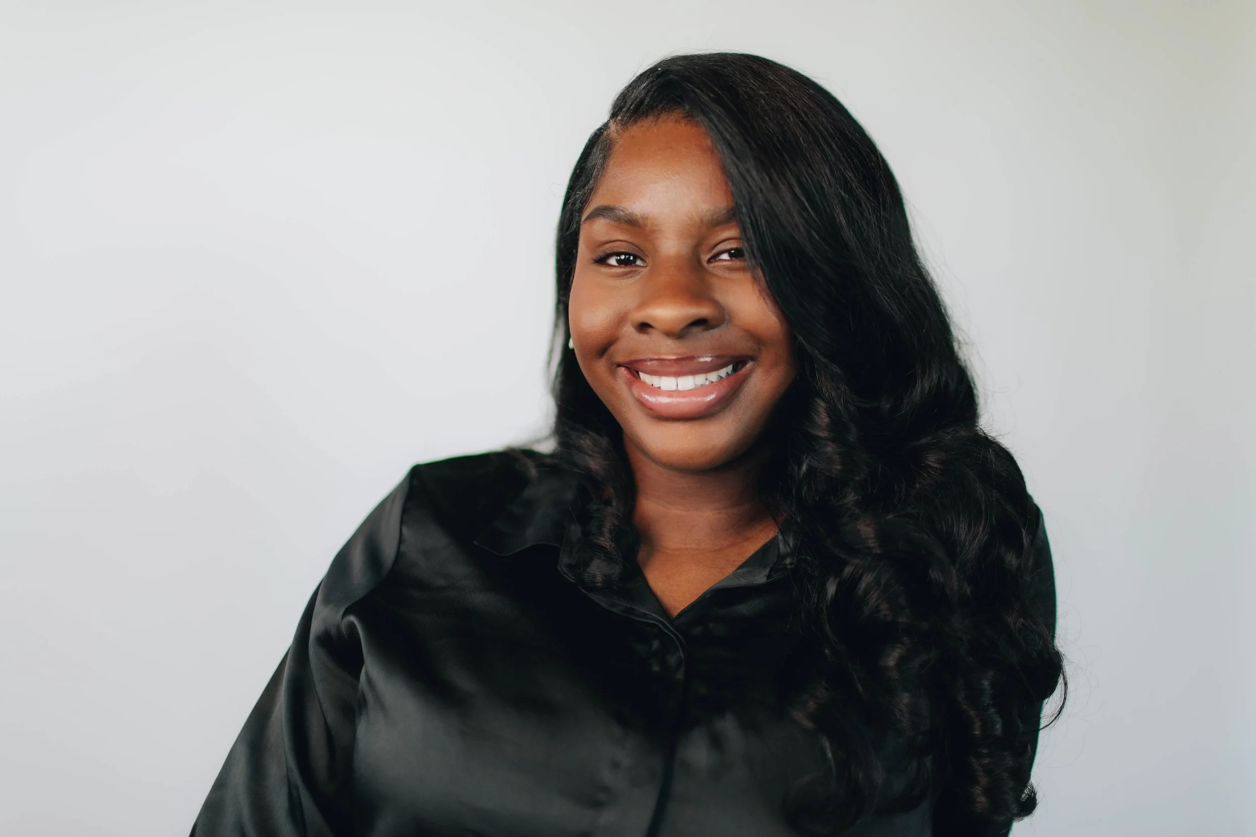 A young woman with dark curly hair smiling, wearing a black satin shirt, against a plain white background.