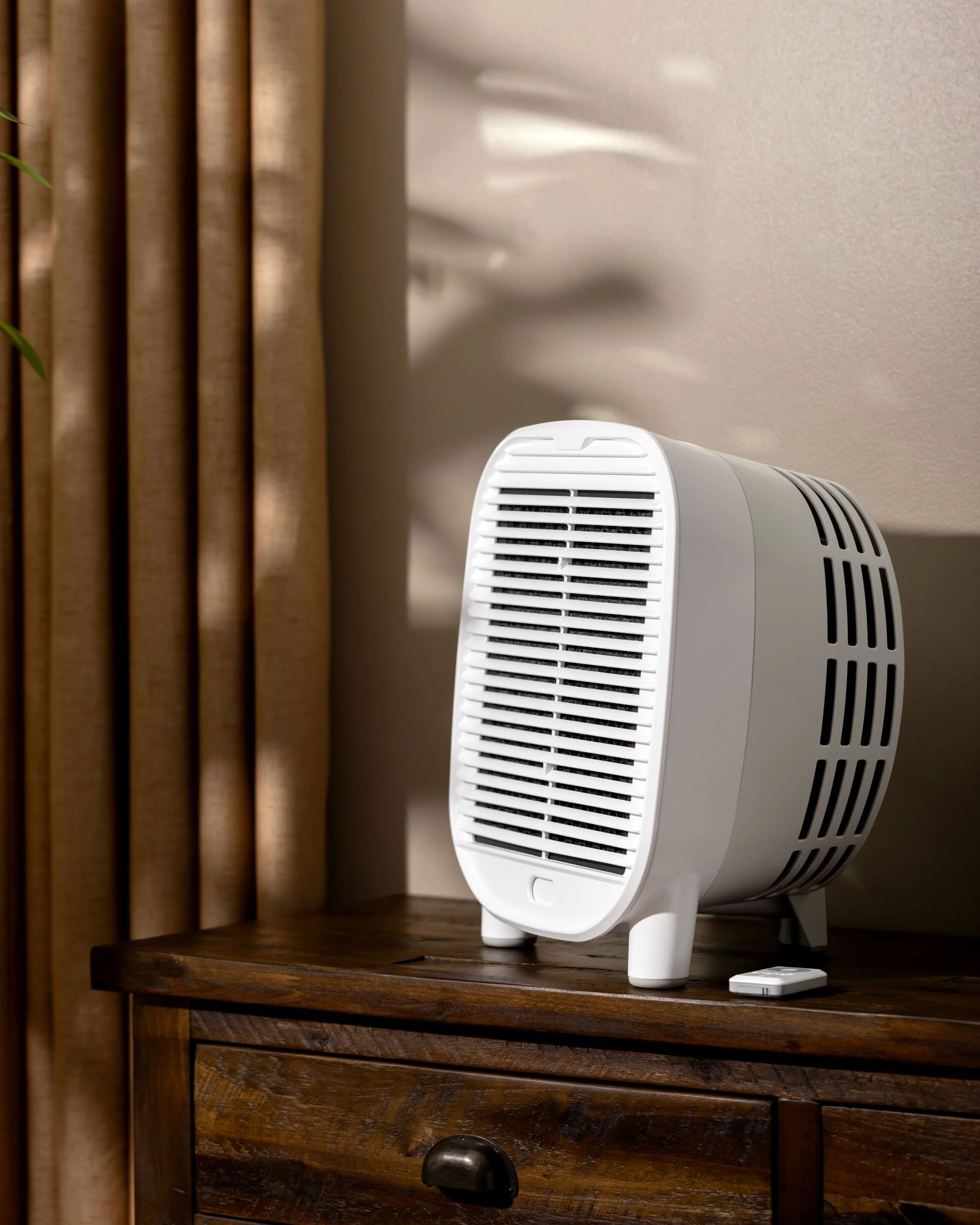 A white space heater with a digital control on a wooden table near beige curtains and a wall.
