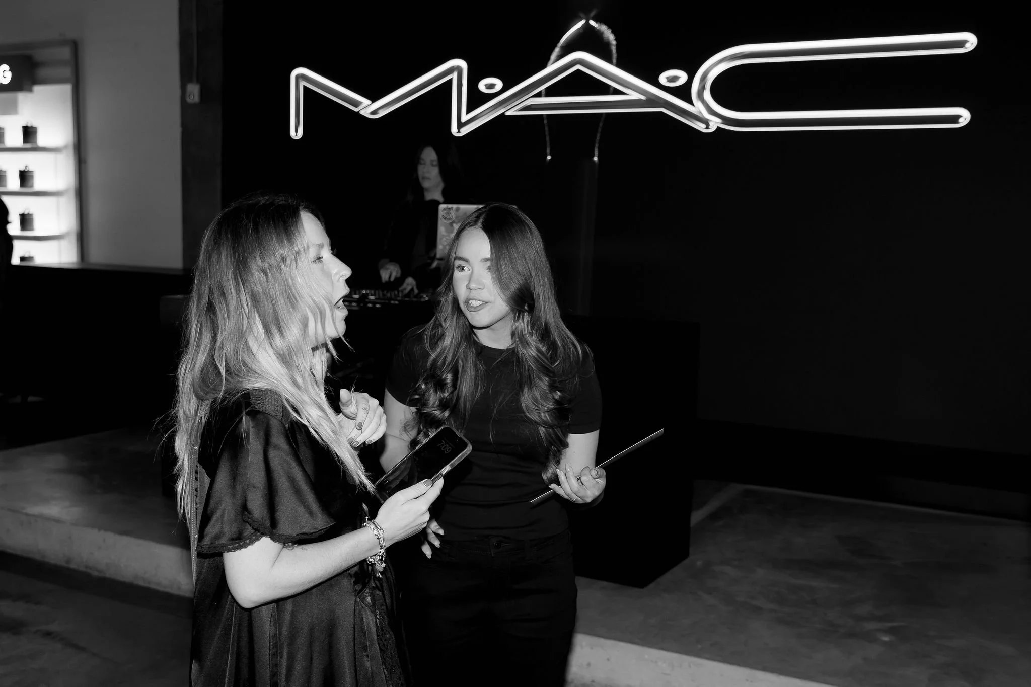 Two women talking in front of a MAC Cosmetics sign. One woman is holding a mobile device and the other is holding a tablet. A DJ is in the background.