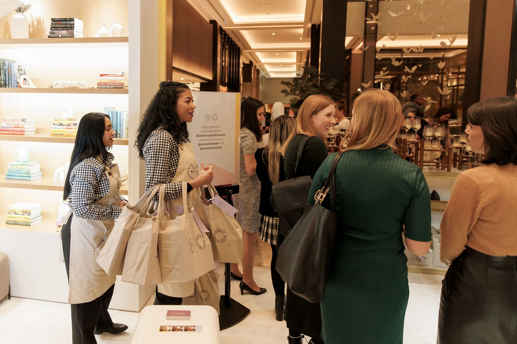 Group of people at a social event in a well-lit room with bookshelves, two women in checkered aprons holding tote bags, and others engaging in conversation.