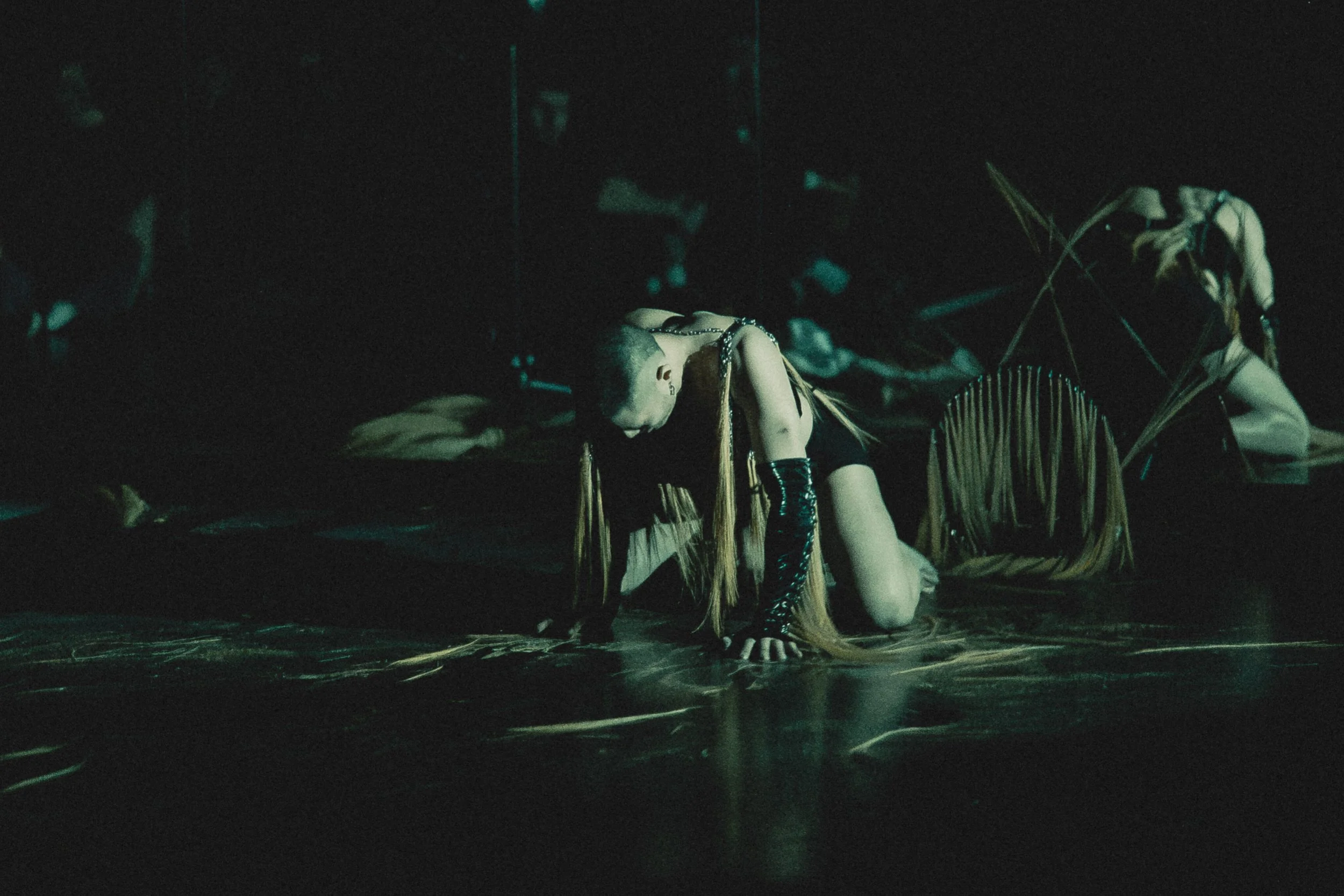 Performer kneeling on the stage with her head bowed down, wearing black outfit with metallic arm warmers, and long hair with extensions, in a dark setting with minimal lighting, possibly during a dance or theatrical performance.