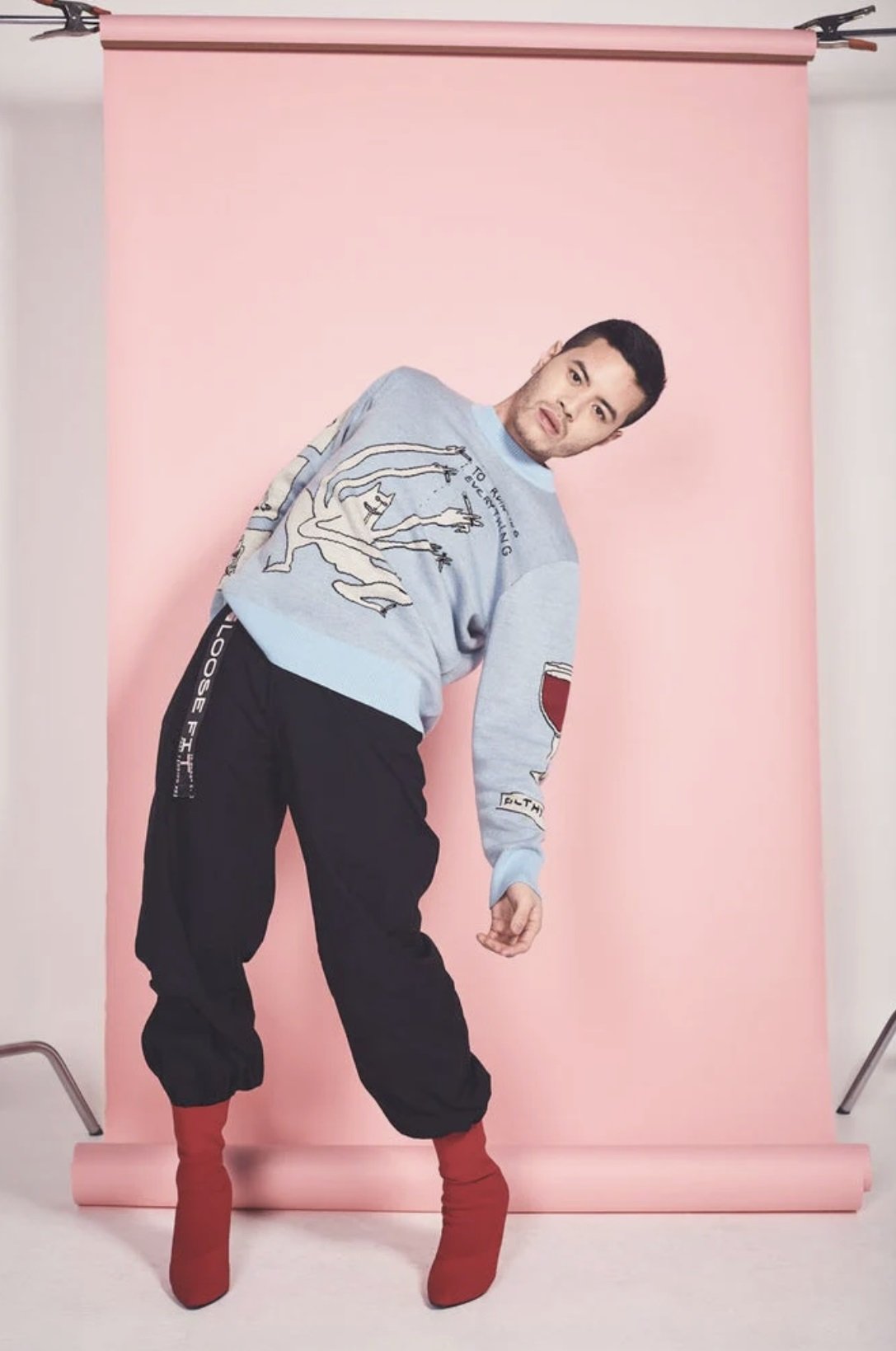 Person in fashionable outfit poses against pink backdrop, wearing a light blue graphic sweater, black trousers, and red high-heeled boots.