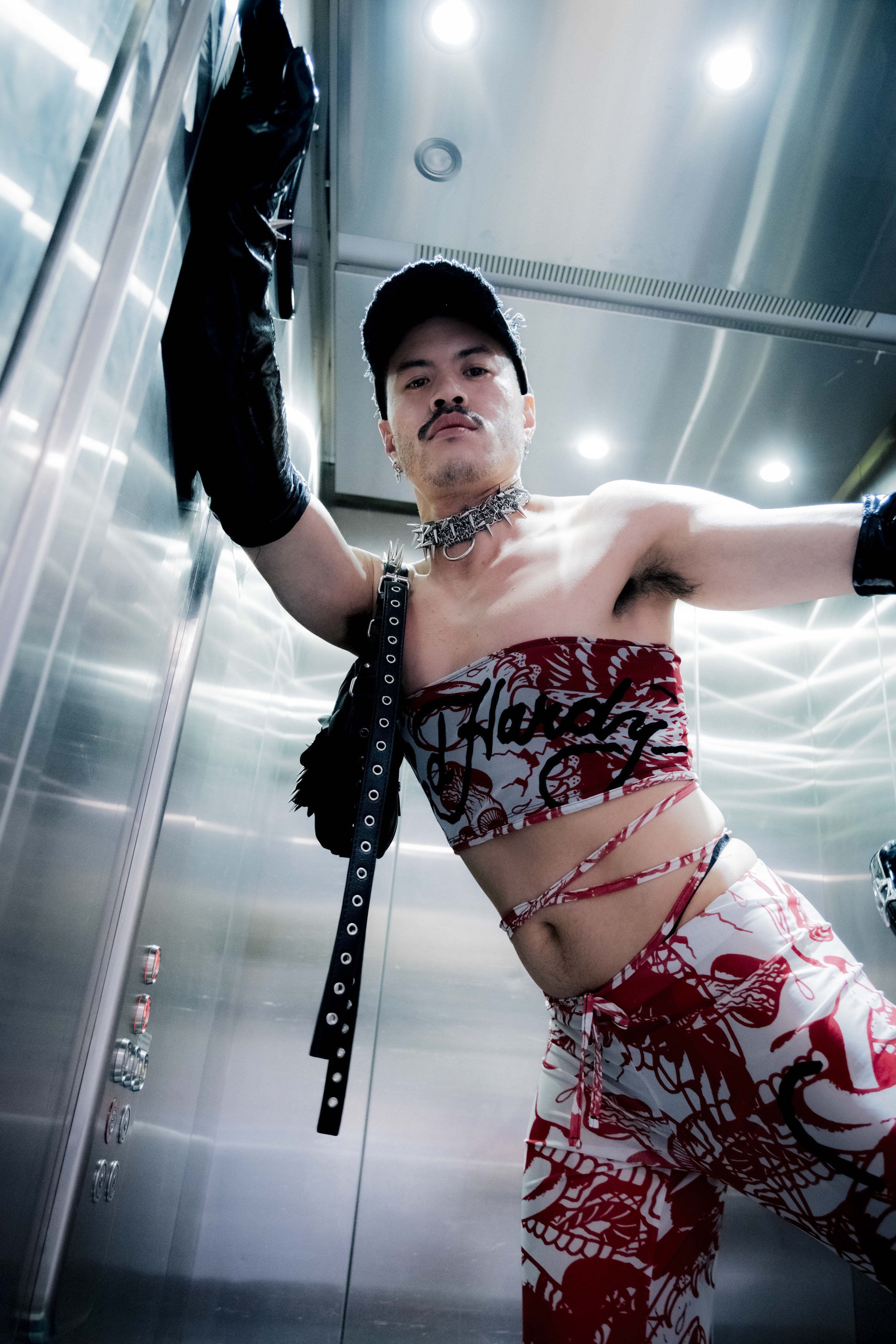 Person in an elevator wearing a red and white outfit with graphical patterns, black gloves, a studded choker, and a cap. The scene is lit by overhead lights.