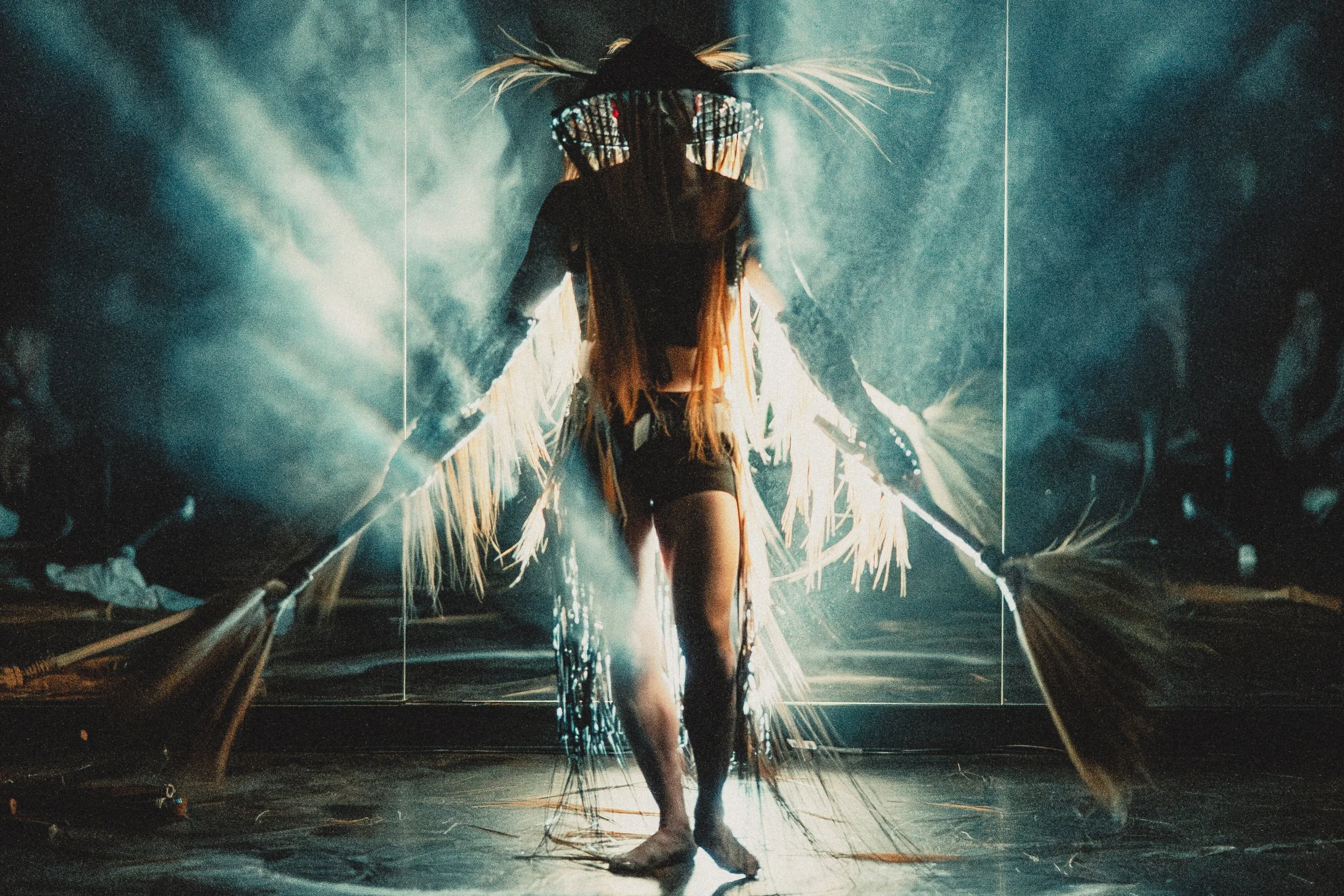 Performer on stage wearing an elaborate costume with fringes, large glasses, and feathers, under dramatic lighting with smoke effects.