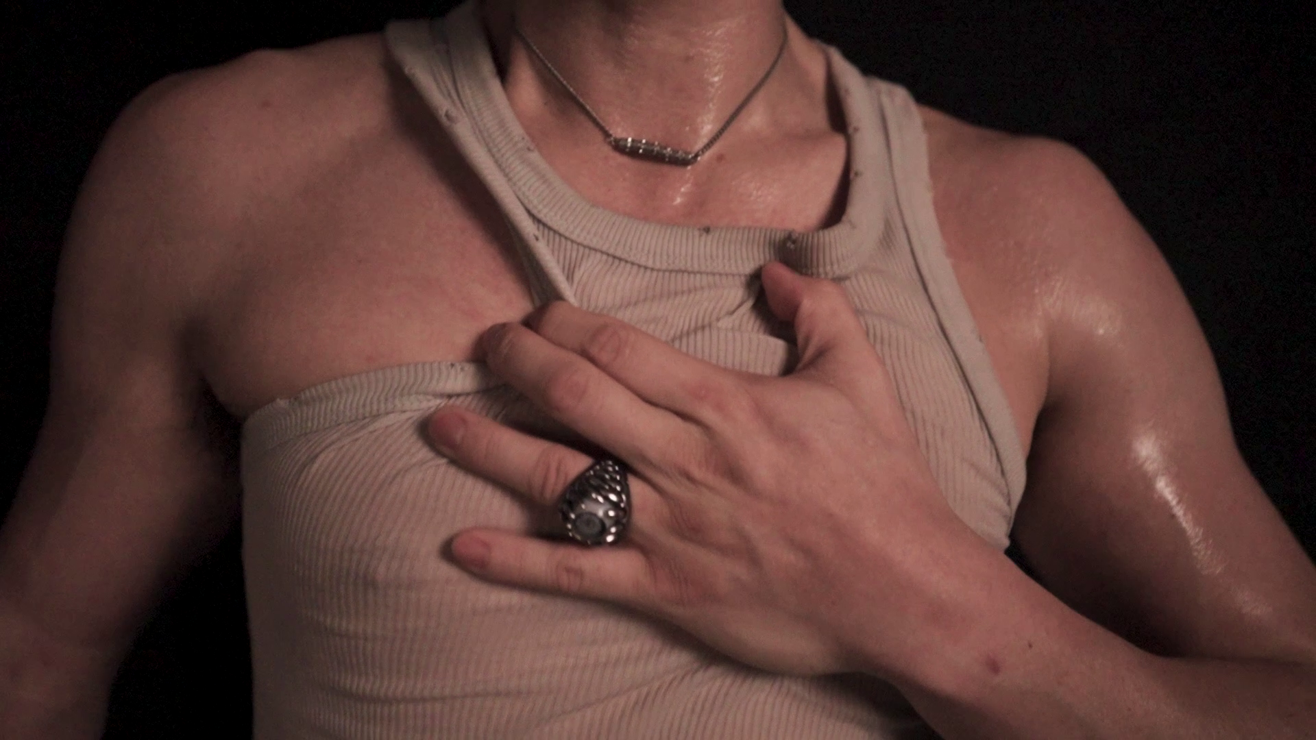 A person wearing a sleeveless shirt, touching their chest with one hand, with visible sweat and a jewelry ring on their finger. Their other hand is partially visible.