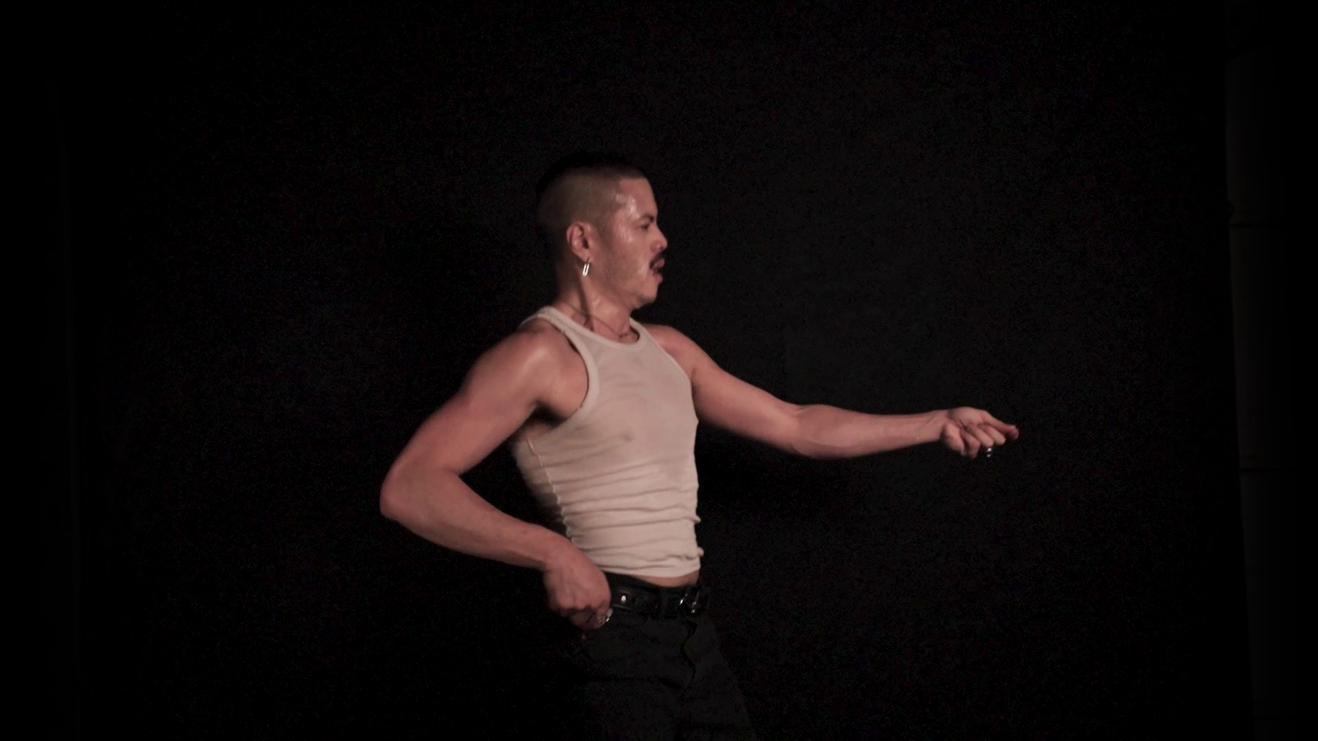 A person with short hair and earrings wearing a beige sleeveless top, standing against a black background, stretching their right arm.