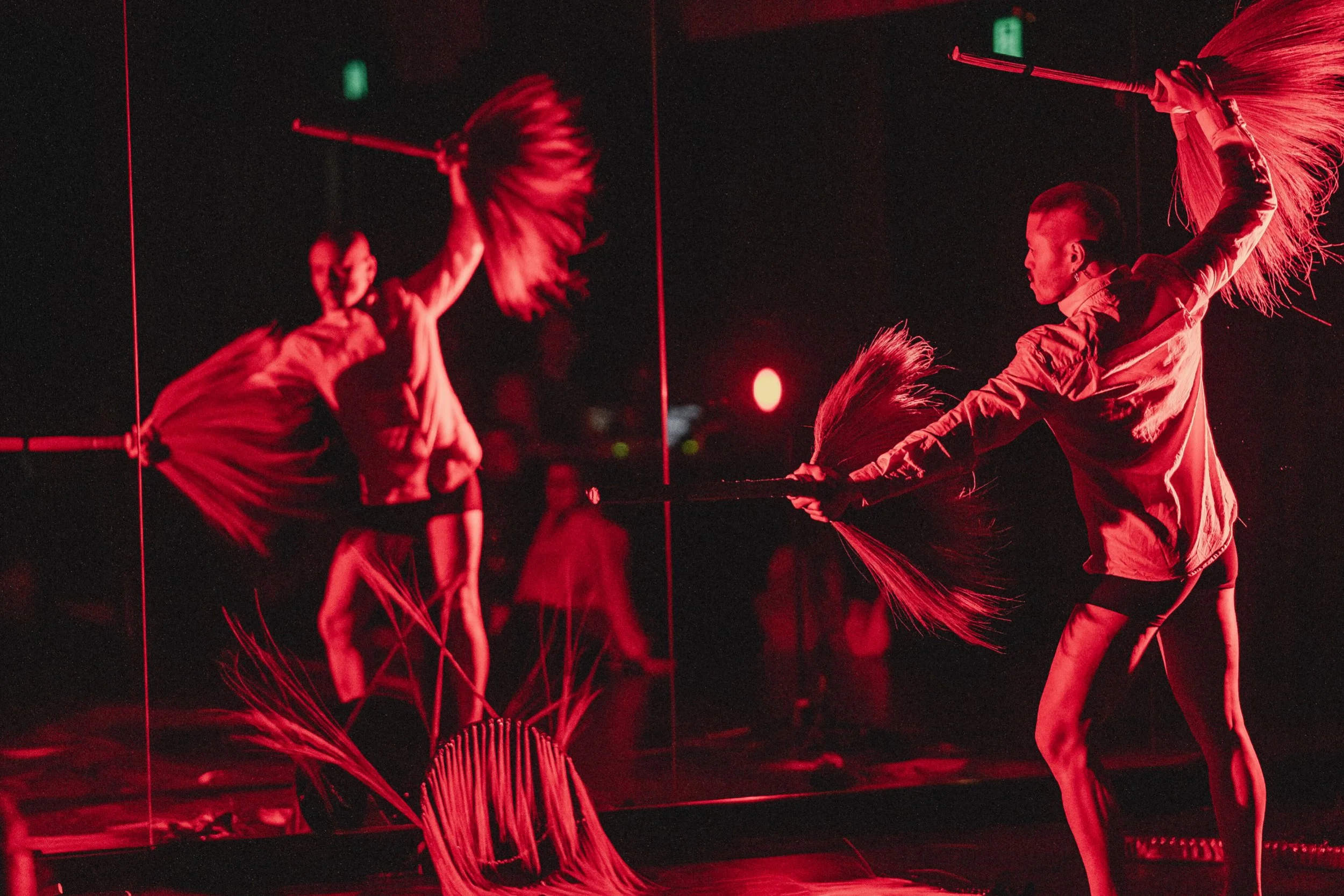 A woman performing a dance or martial arts move, swinging a long stick with tassels, reflected in a mirror in a dimly lit space with red lighting, with some onlookers in the background.