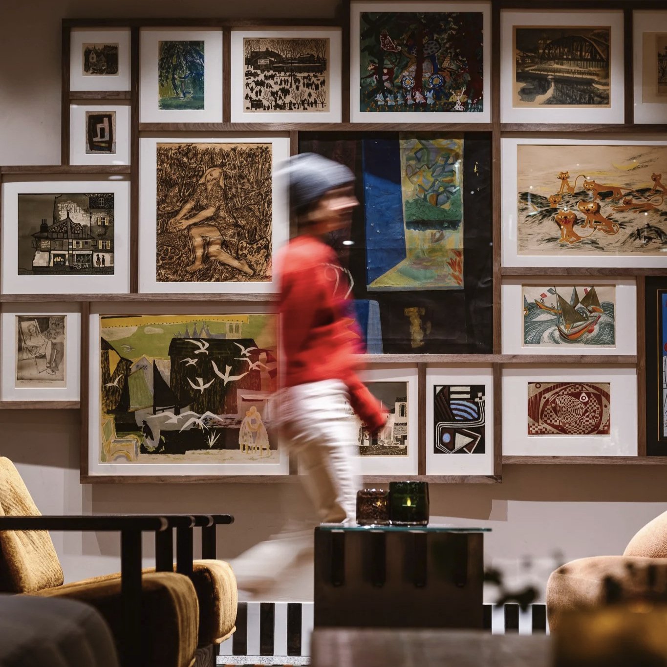 Modern indoor seating area with wooden lounge chairs, blue and white cushions, and a blue bar with decorative canopies. Large windows on the right and overhead lighting.