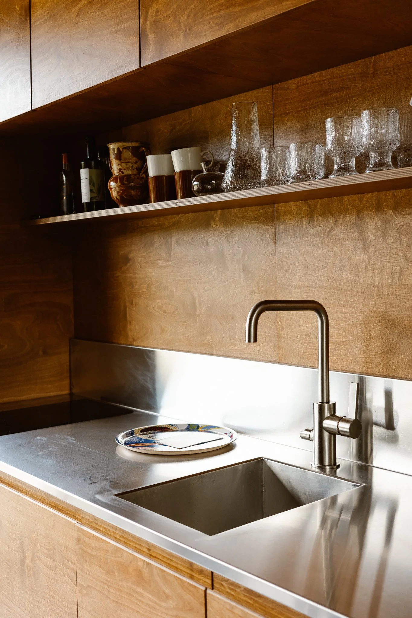 A modern kitchen sink area with a stainless steel sink, wooden cabinets, and open shelves with glassware and bottles.