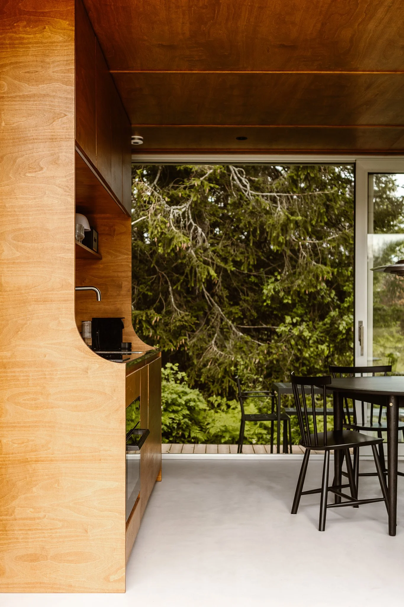 Interior view of a modern kitchen overlooking an outdoor green landscape with trees and a deck, featuring wooden cabinetry and black chairs around a table.
