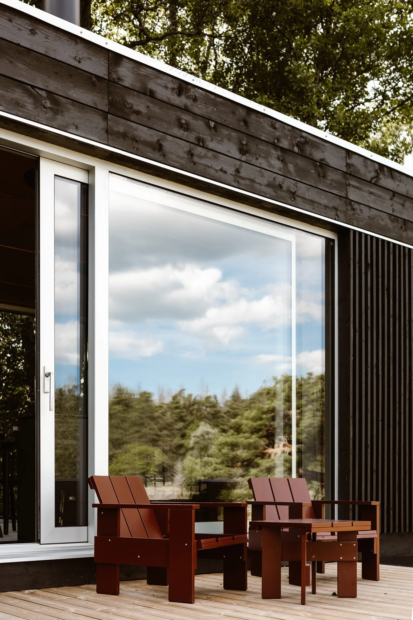 Outdoor patio with two red wooden chairs and a matching side table in front of a large glass window, with a reflection of blue sky and white clouds, surrounded by trees.