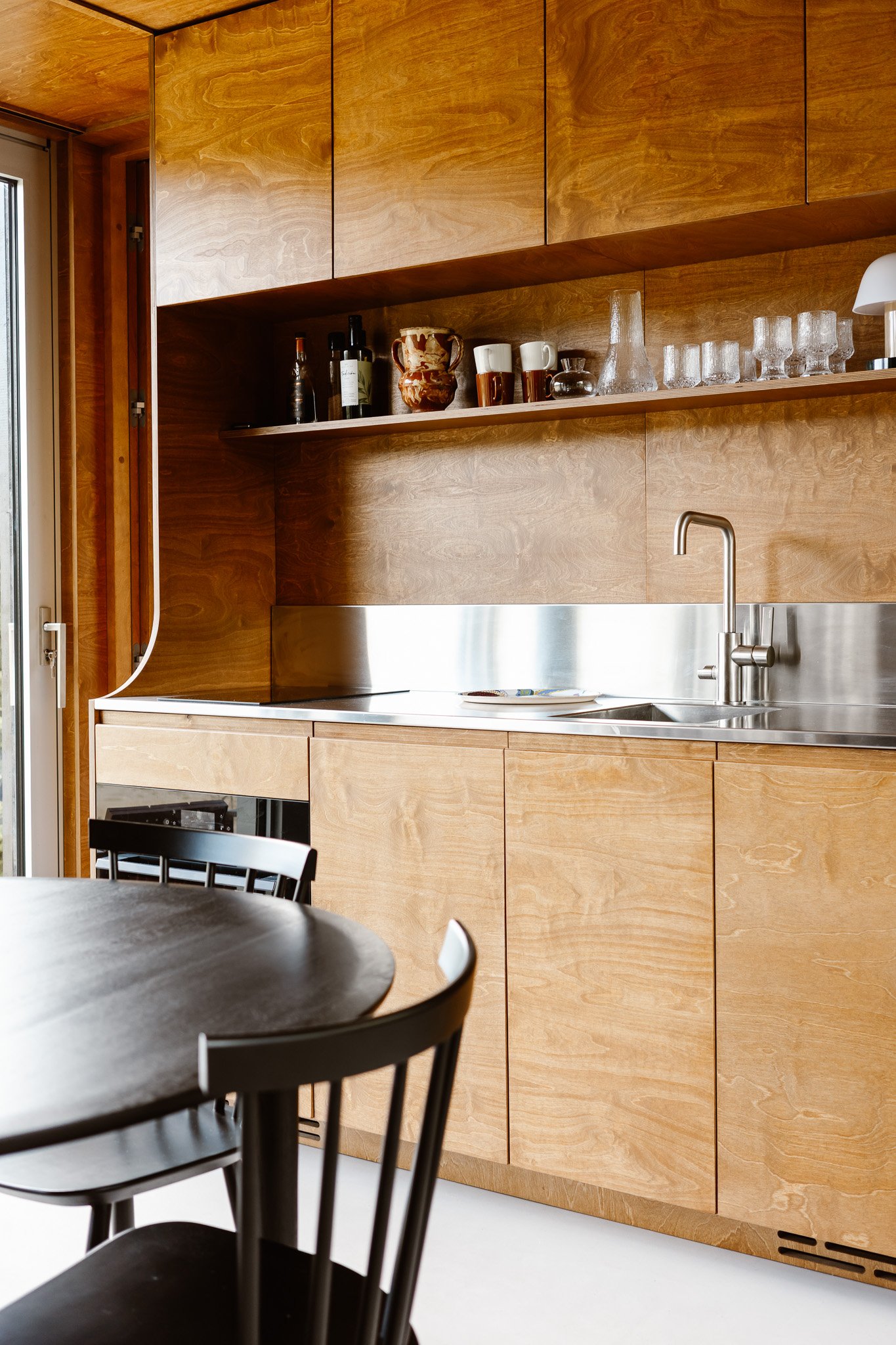 A modern kitchen with wooden cabinets, a stainless steel countertop and backsplash, a sink with a faucet, a shelf with drinking glasses and decorative items, a window to the left, and a black chair at a round table in the foreground.