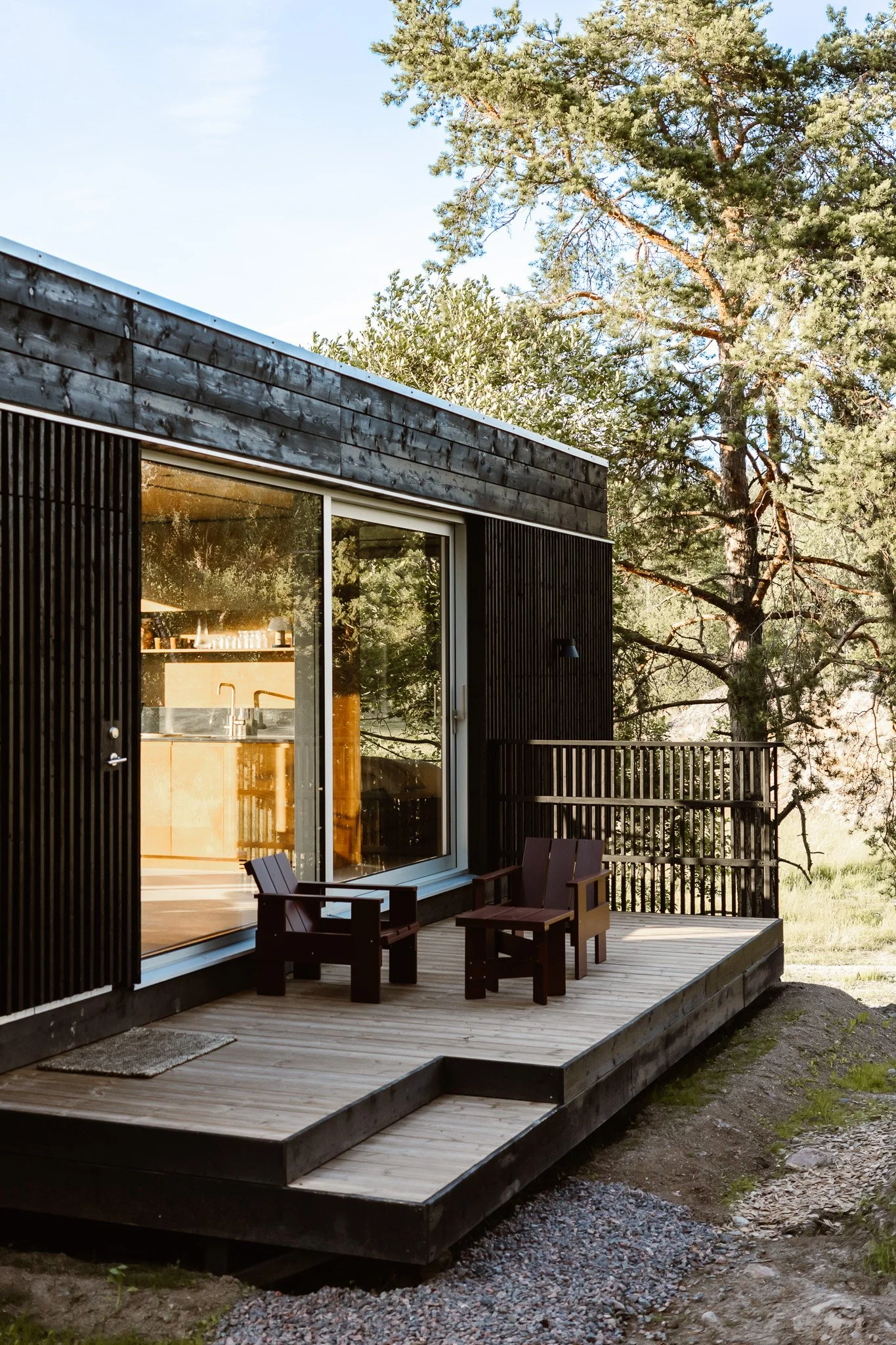 A modern black wooden house with a glass sliding door and a small outdoor wooden deck with two Adirondack chairs, set in a wooded area with tall trees.