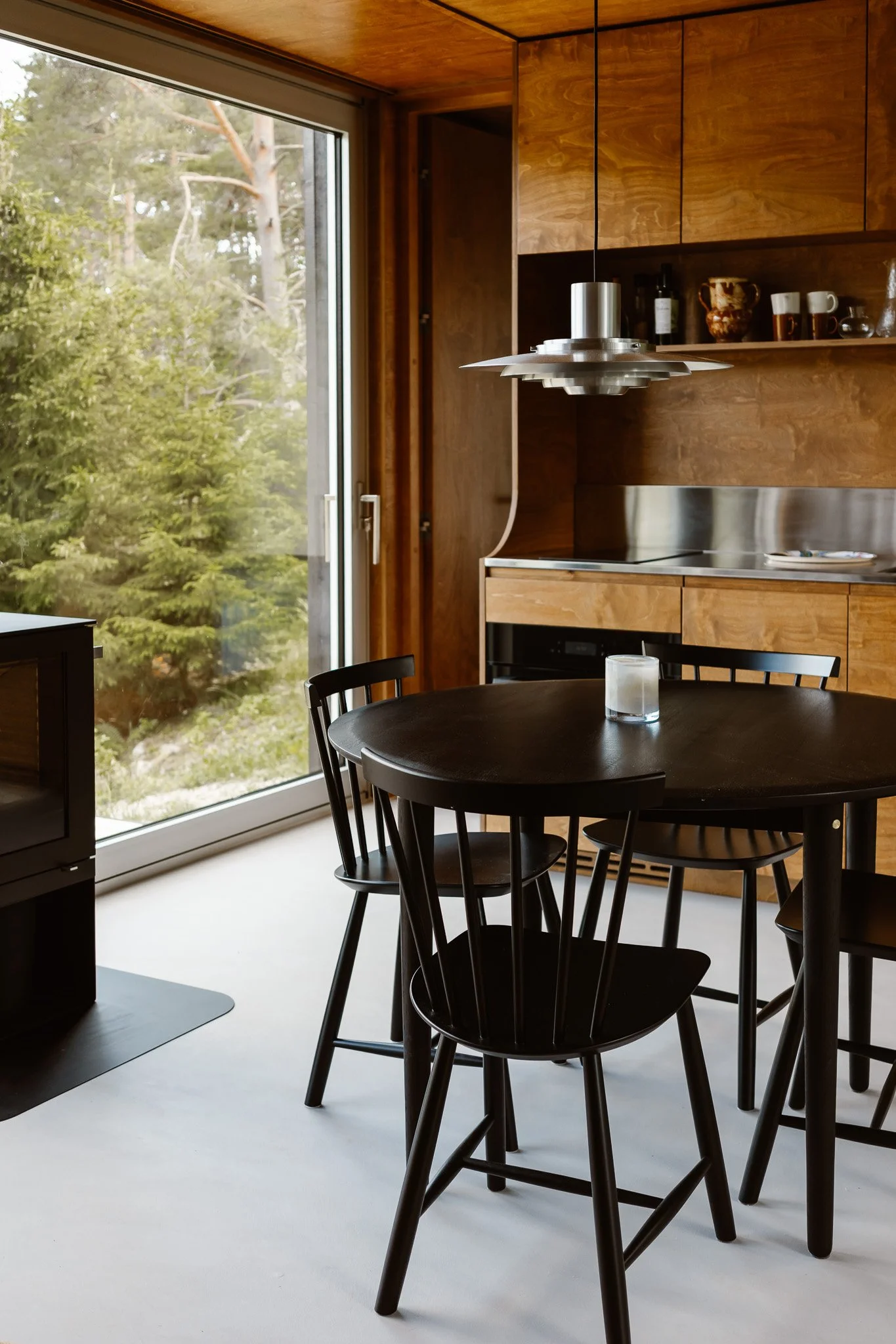 A kitchen with wooden cabinets, a black dining table with four black chairs, a sliding glass door leading to greenery outside, and a modern hanging light fixture.