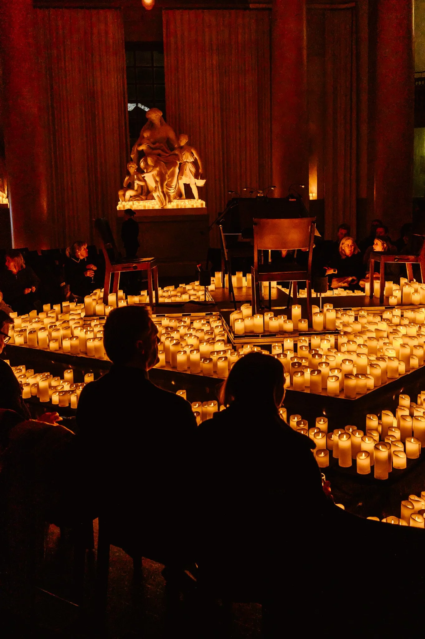 Interior scene with numerous candles and people seated around a central arrangement, in front of a statue.