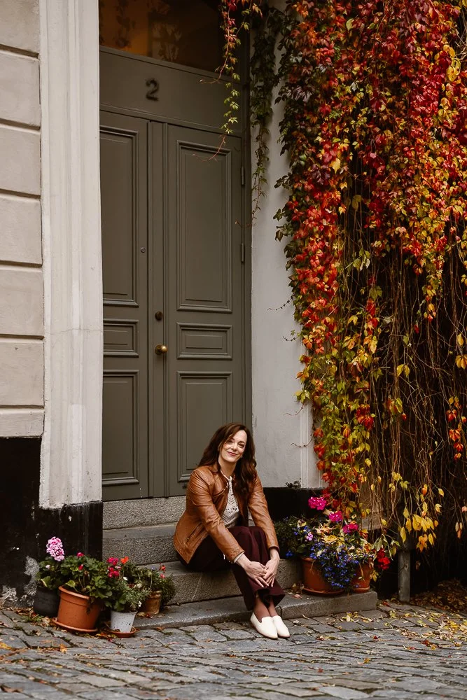 A woman with long brown hair, wearing a brown leather jacket, white top, dark pants, and white shoes, sitting on stone steps in front of a green door numbered 2. Surrounding her are pots of colorful flowers and a wall covered with red, orange, and ye