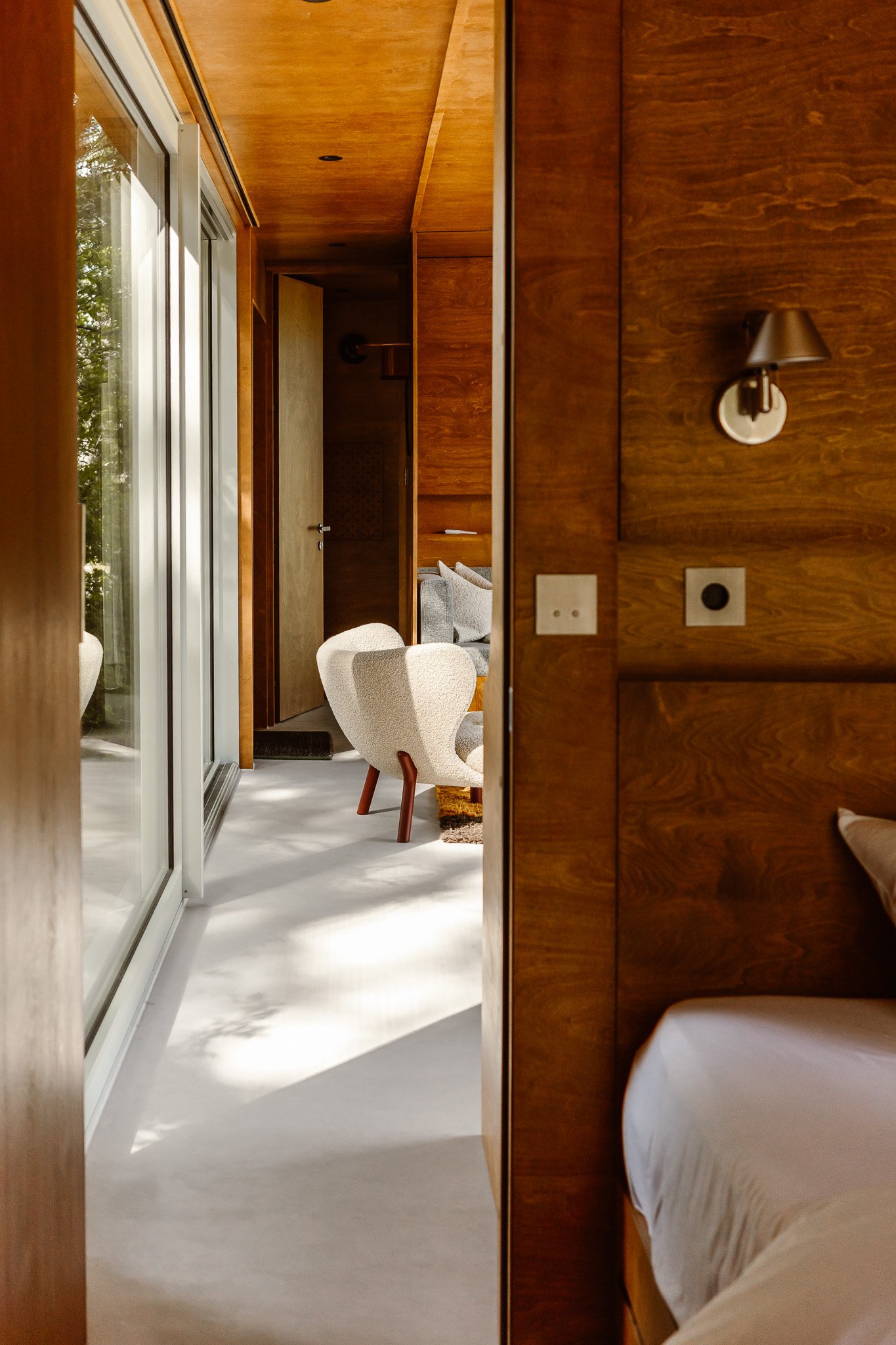 Interior view of a living space with wood-paneled walls, a bedroom on the right, and a sitting area with a beige armchair and a sofa in the background, with sunlight filtering through sliding glass doors.