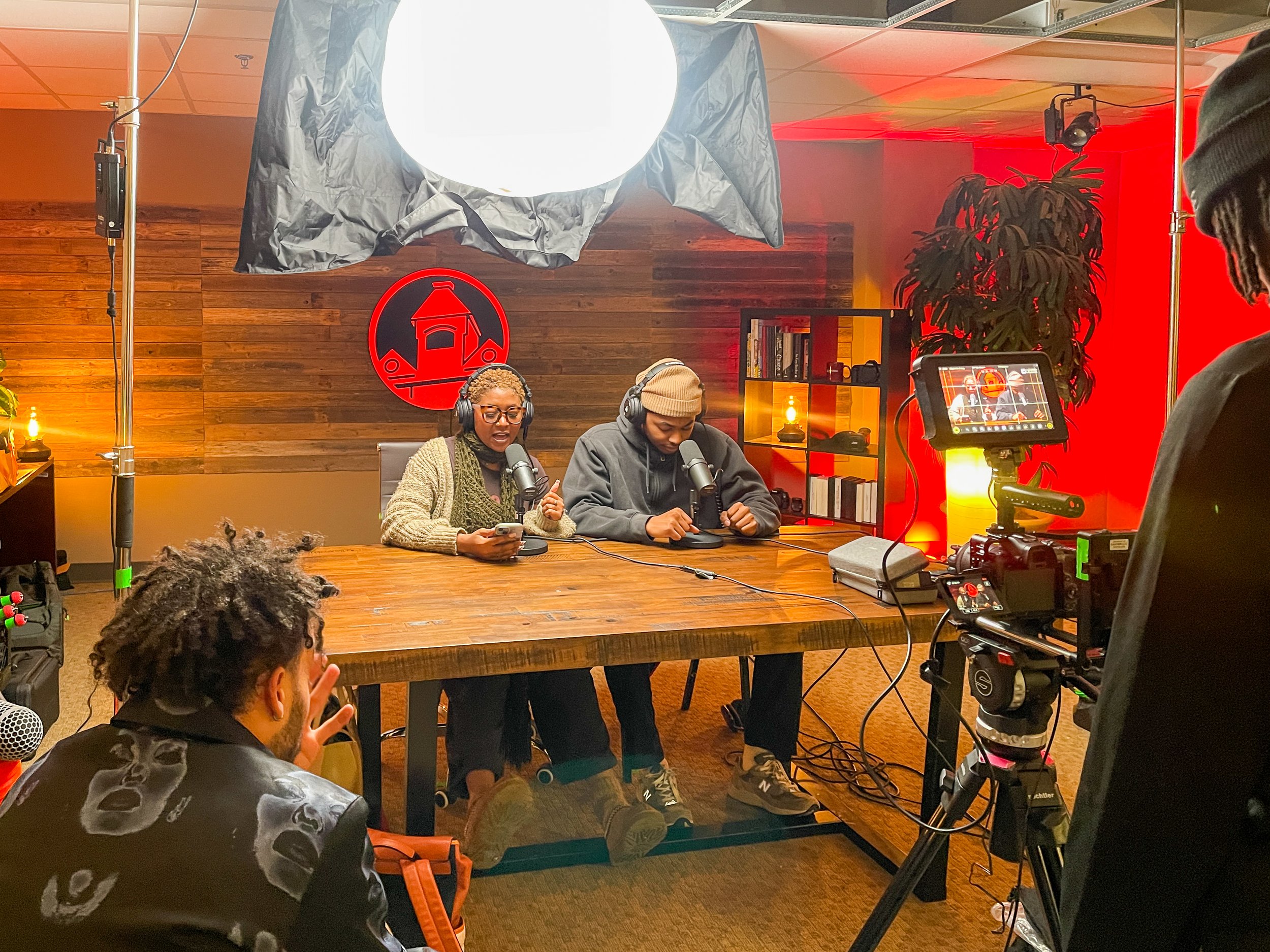 Podcast recording setup with two hosts and camera crew, featuring microphones, lighting, and a wooden table in a studio with red accents.