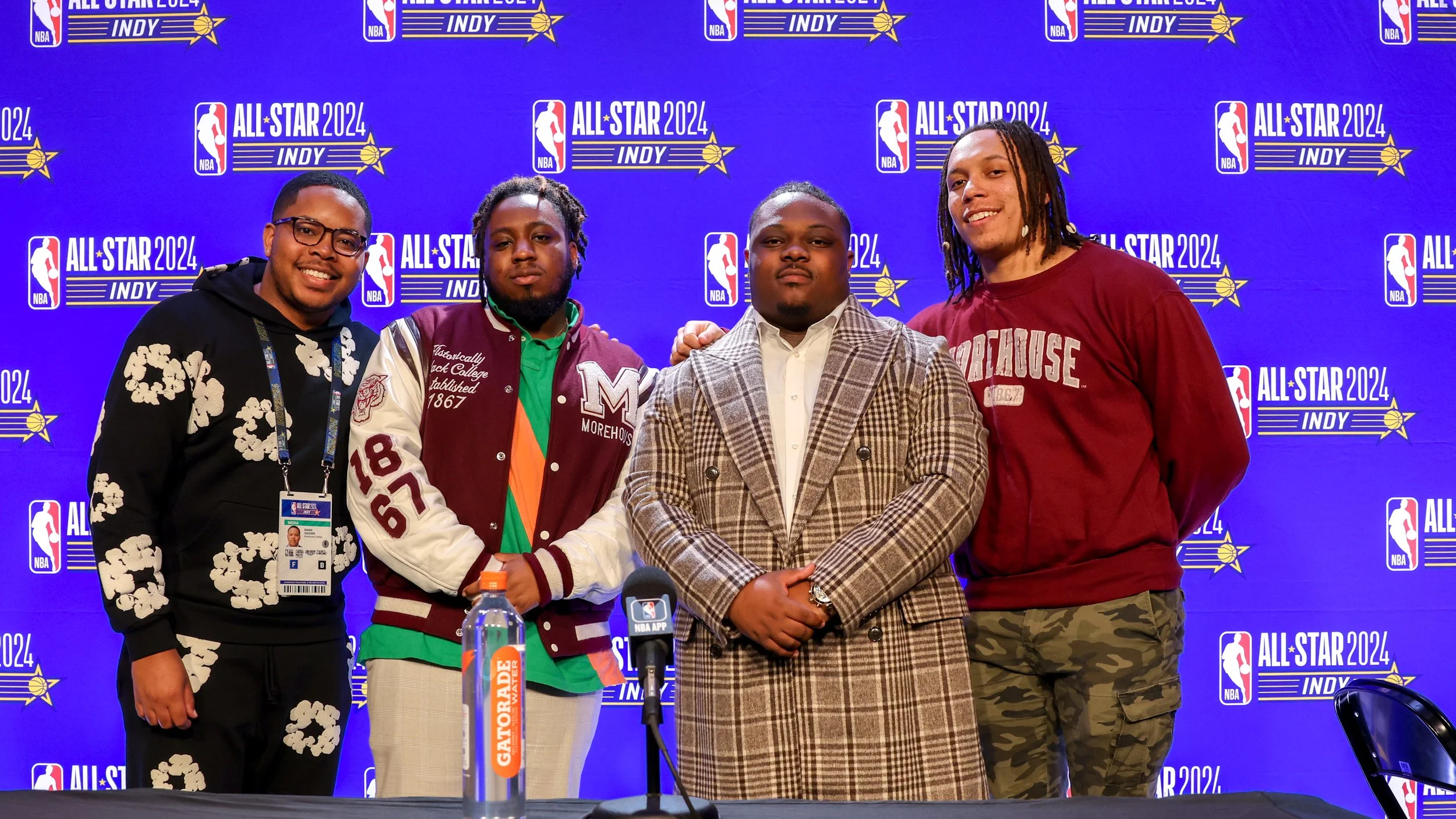 Four men standing together in front of a backdrop for the NBA All-Star 2024 event in Indianapolis. One is wearing a flower-patterned hoodie, another a Morehouse jacket, a third a checked coat, and the fourth a Morehouse sweatshirt. A Gatorade bottle 