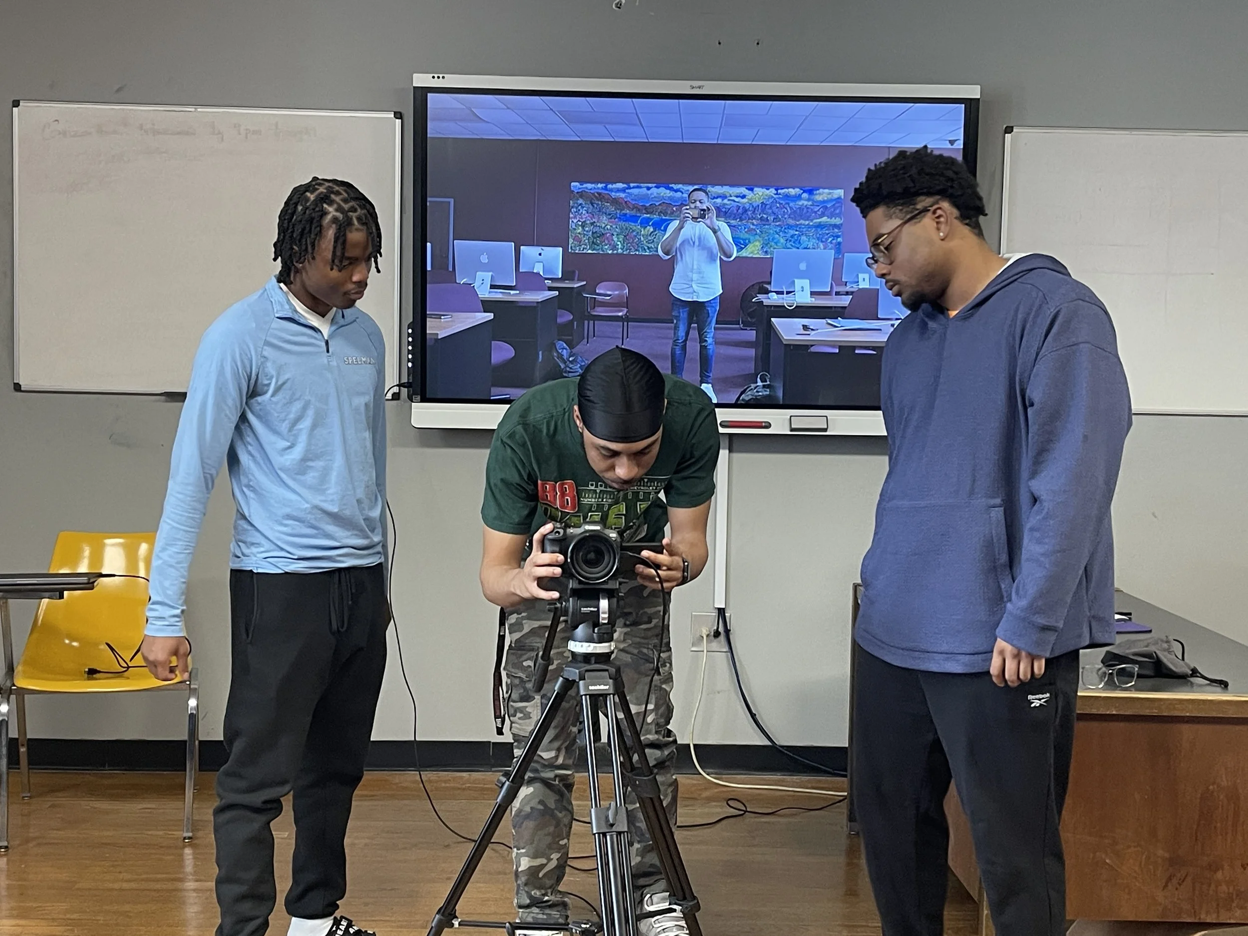 Three people in a classroom setting, one operating a camera on a tripod, with others observing. A digital screen in the background shows a mirrored image of the cameraman.