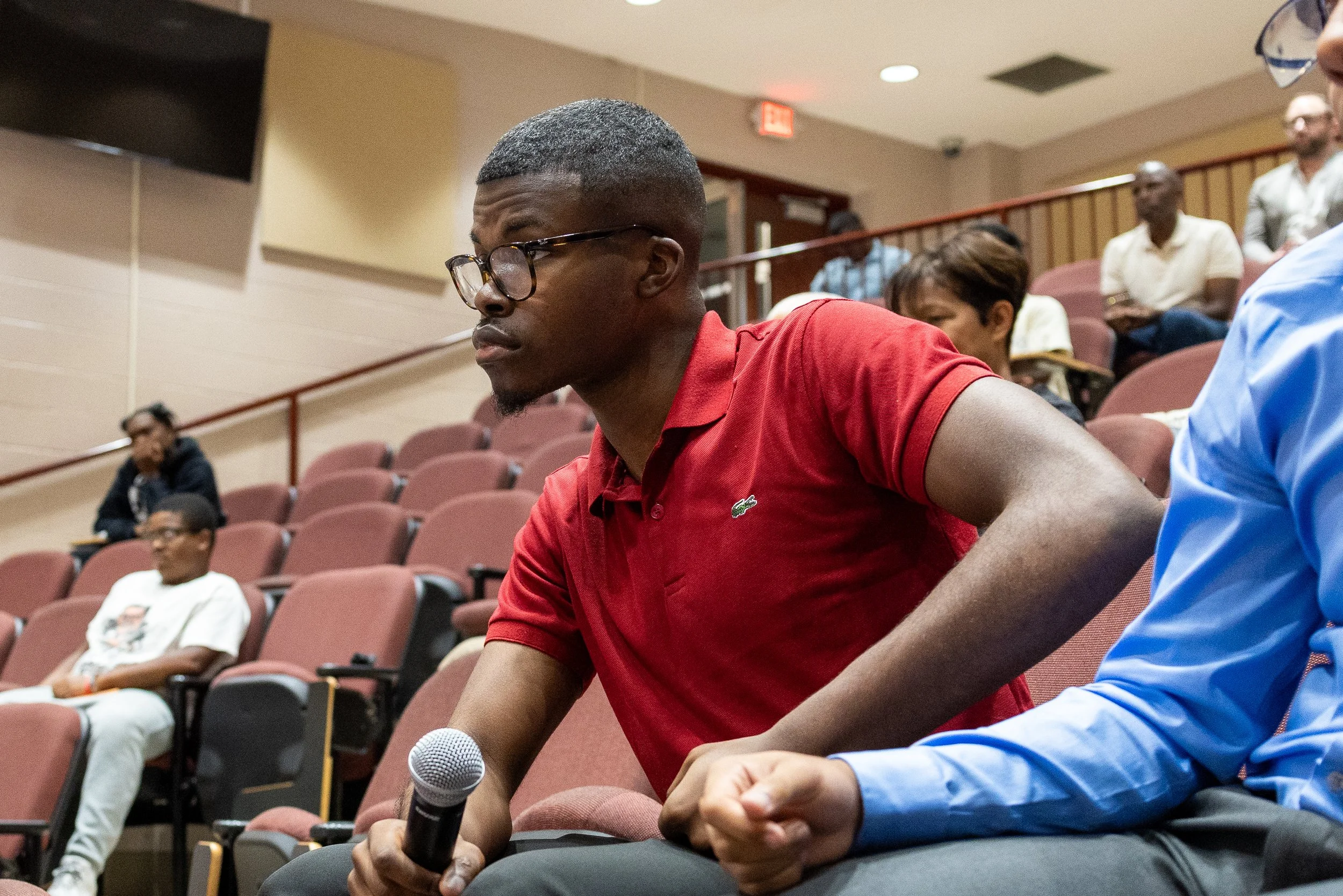 Person in a red shirt holding a microphone seated in an auditorium with other people.