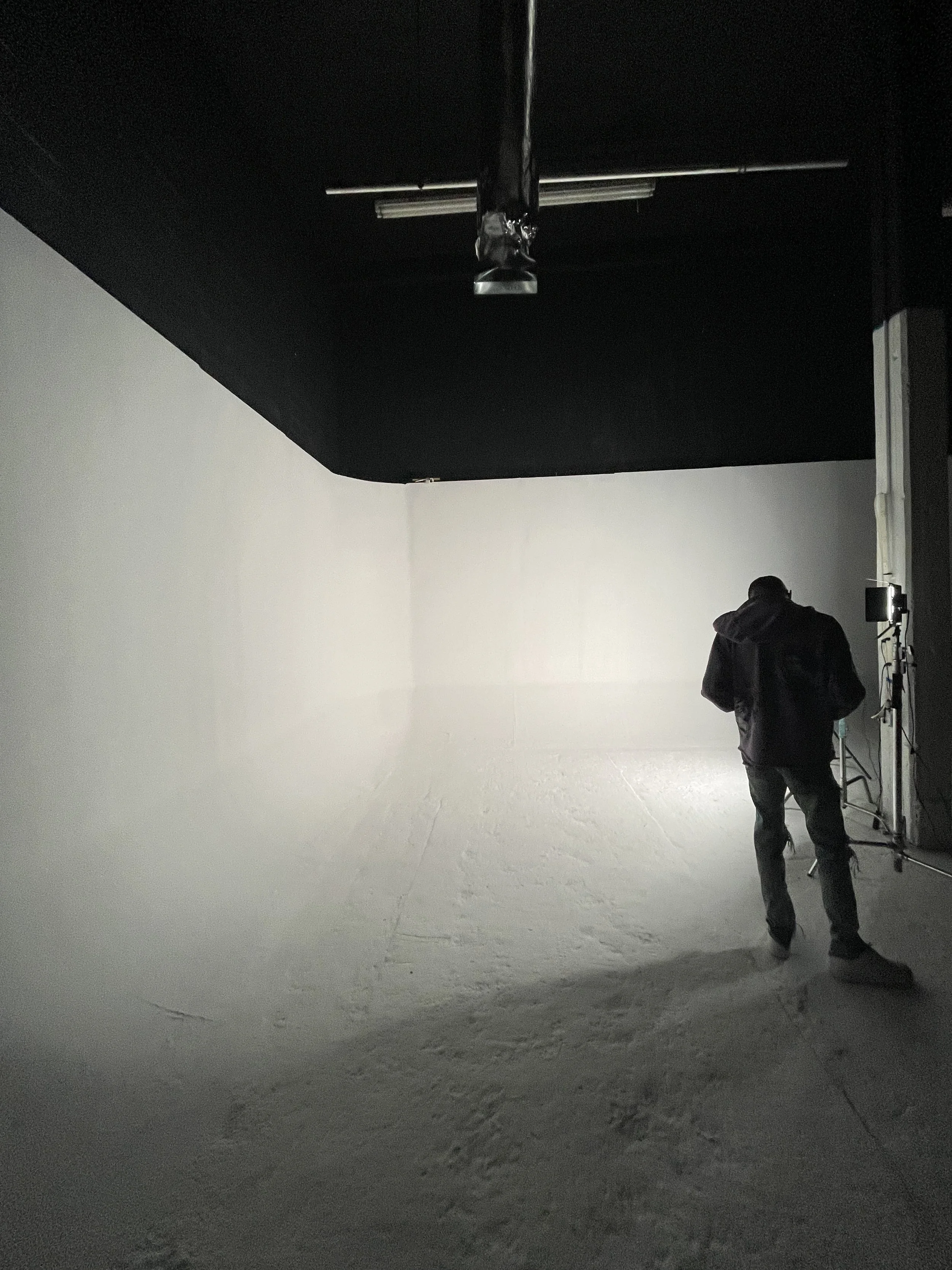 Person standing in a dimly lit room with white walls and floor, possibly a photography studio with equipment.