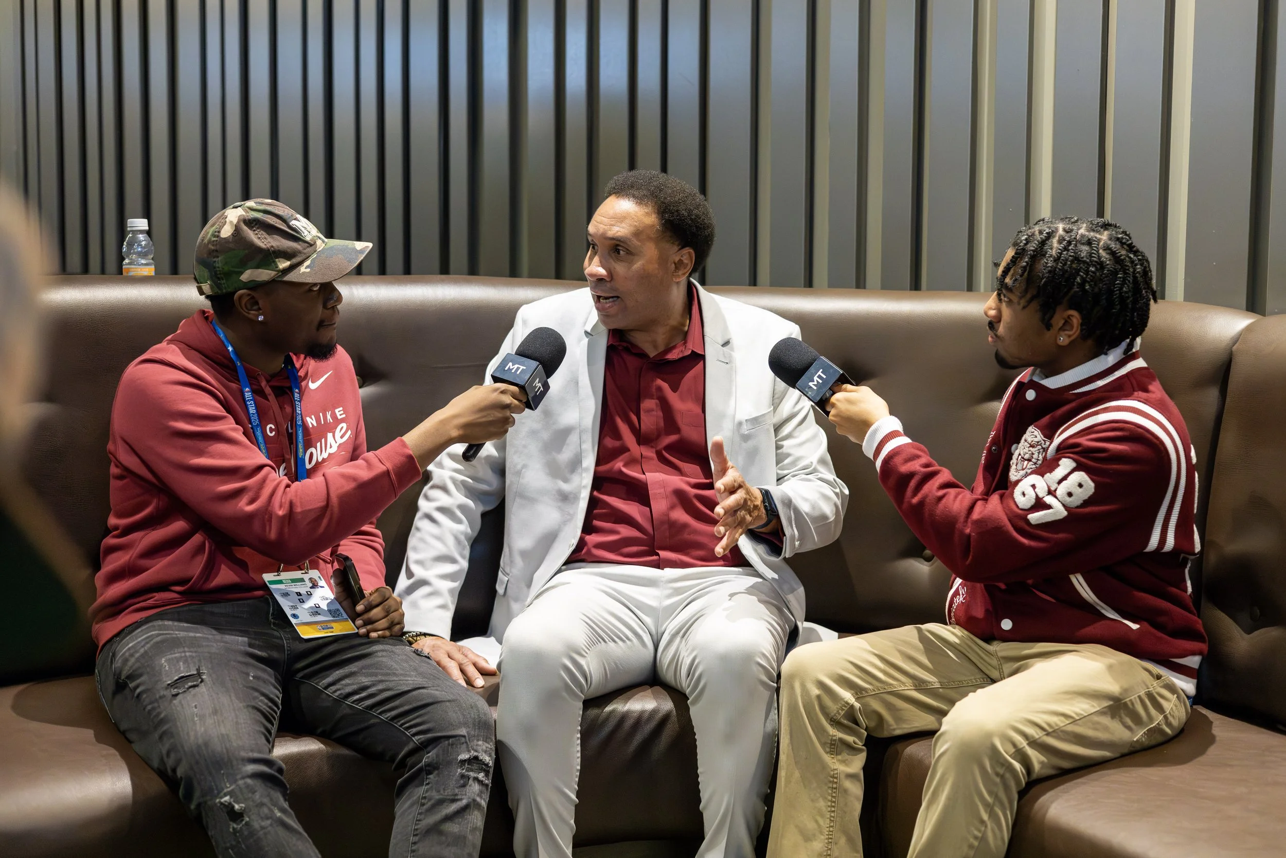 Three men sitting on a couch having a conversation. Two are holding microphones, interviewing the man in the middle. The man in the center is wearing a white suit and speaking. The other two are in casual attire, one wearing a red jacket and the othe