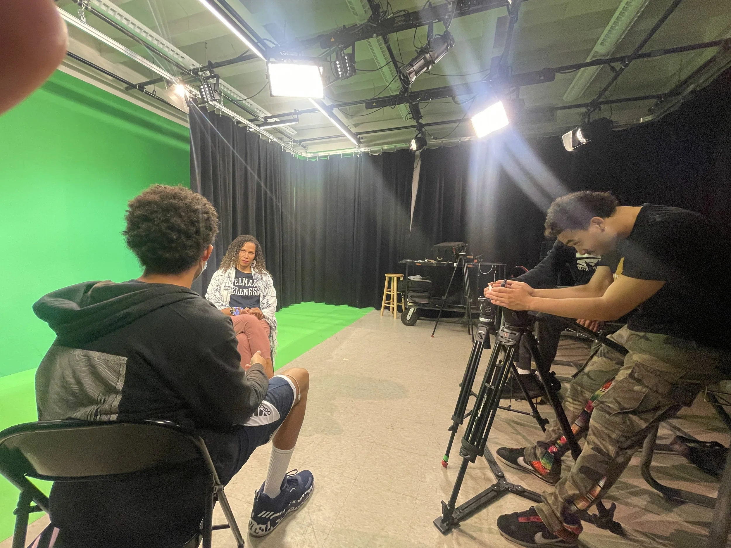 People in a studio with green screen, camera setup, and lighting equipment. One person is seated being interviewed, another adjusting a camera.