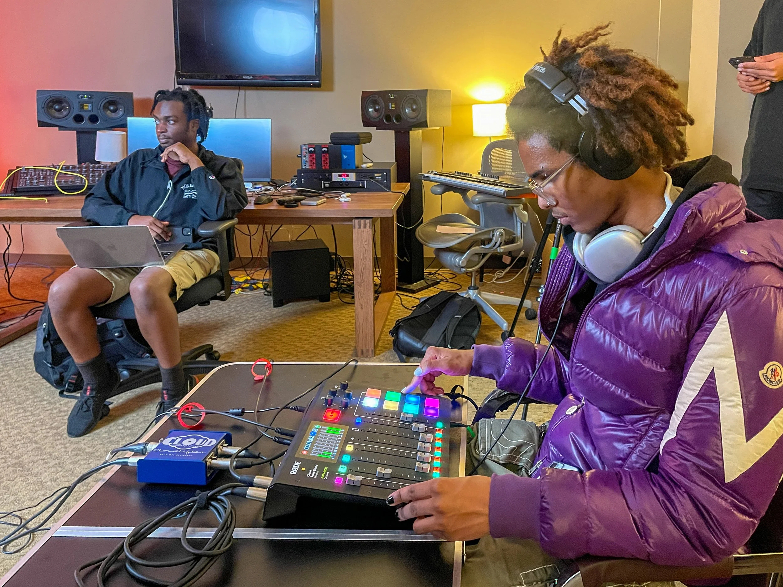 Two people in a recording studio, one using audio equipment and the other with a laptop, surrounded by sound gear.