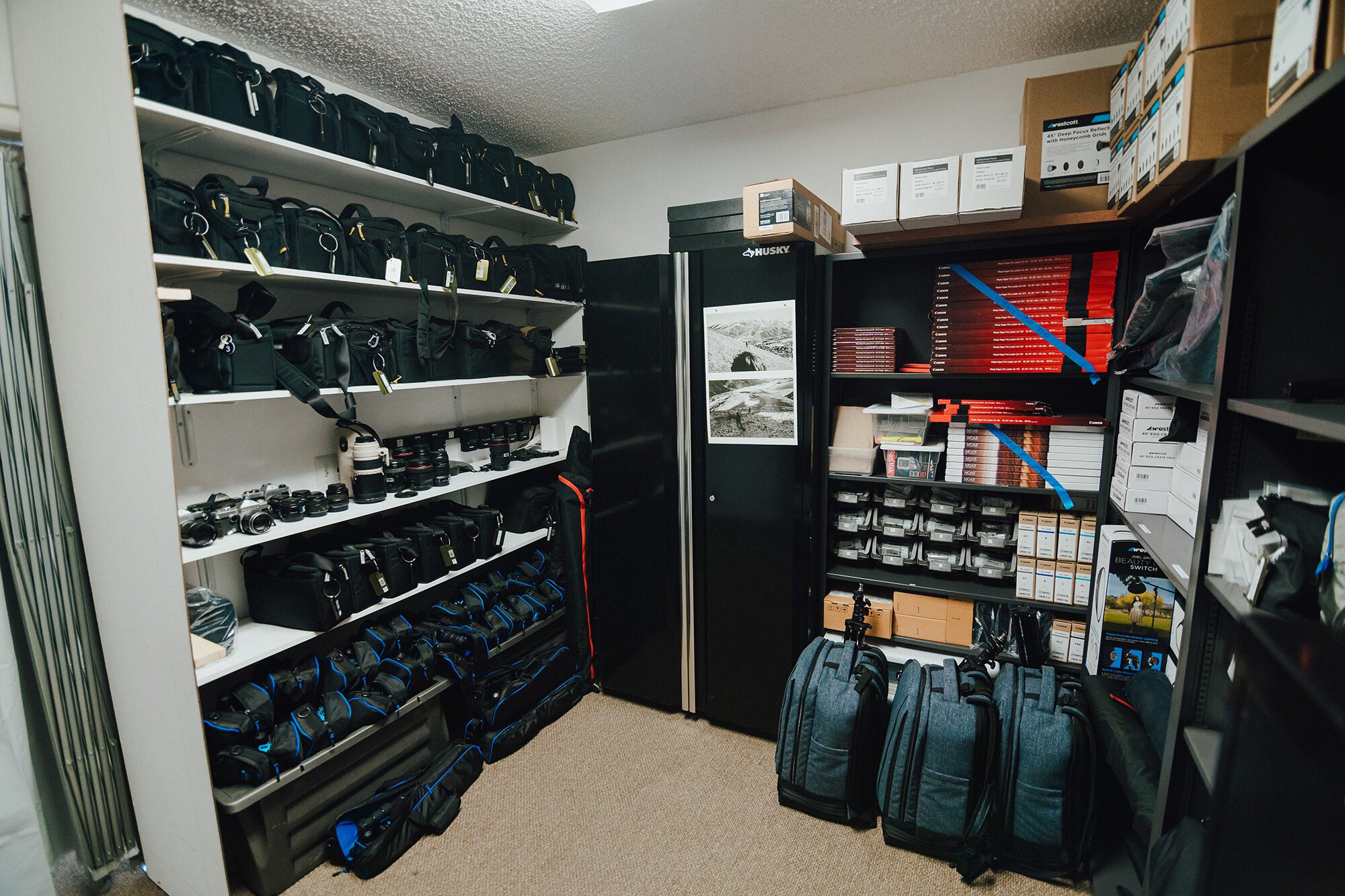 Room with shelves of camera bags, lenses, equipment boxes, and storage cabinets.