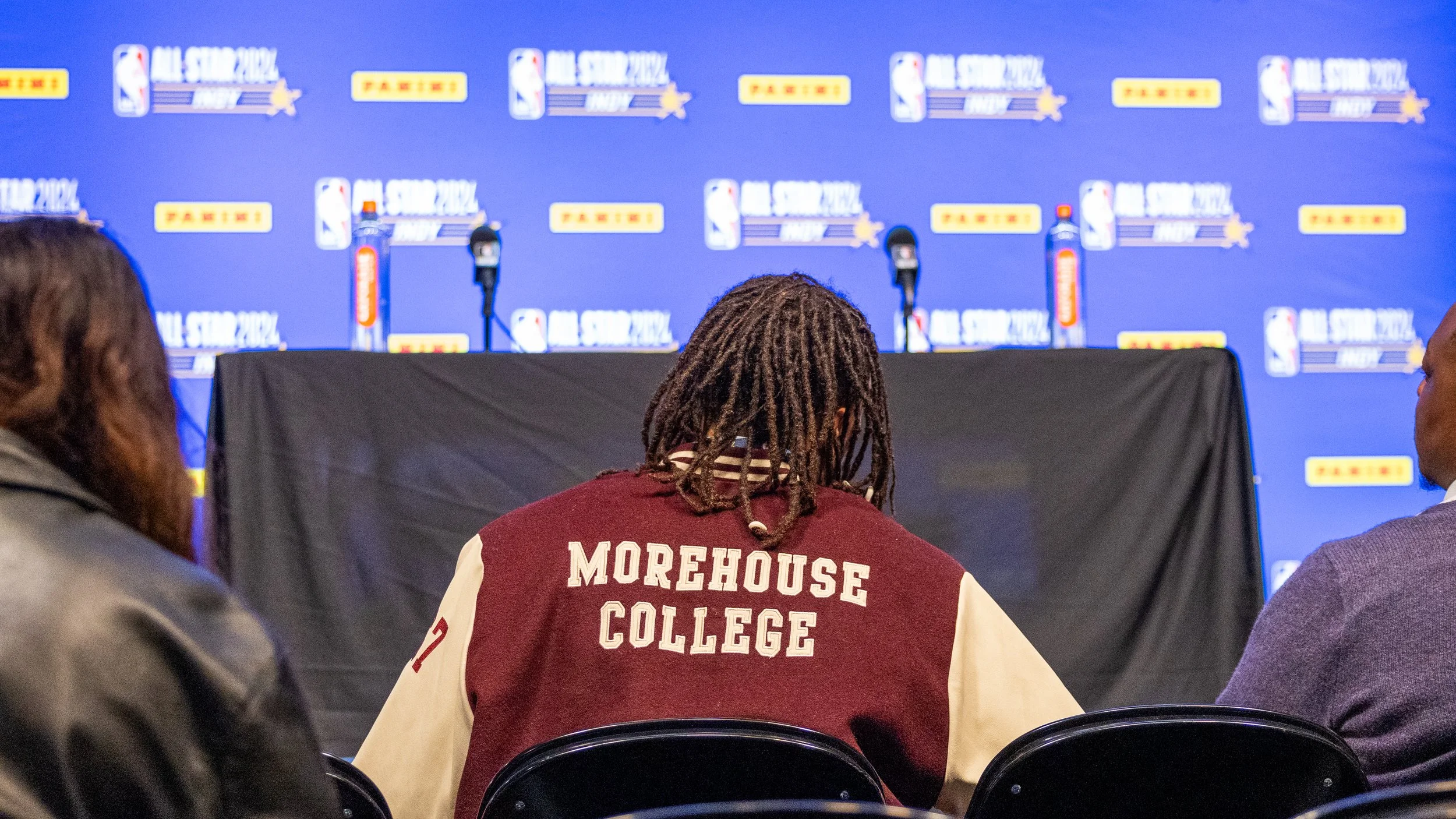 Person wearing Morehouse College jacket at NBA All-Star 2024 press event with microphones and backdrop logos.