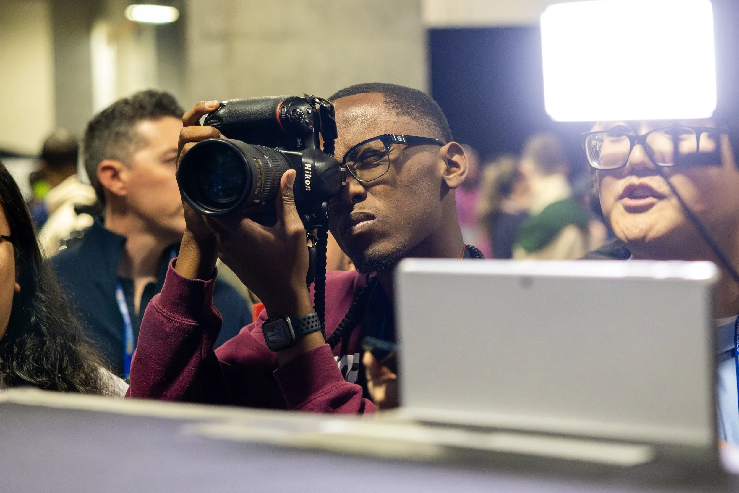 A person holding a Nikon camera and taking photos in a crowded indoor environment, with other people visible in the background.
