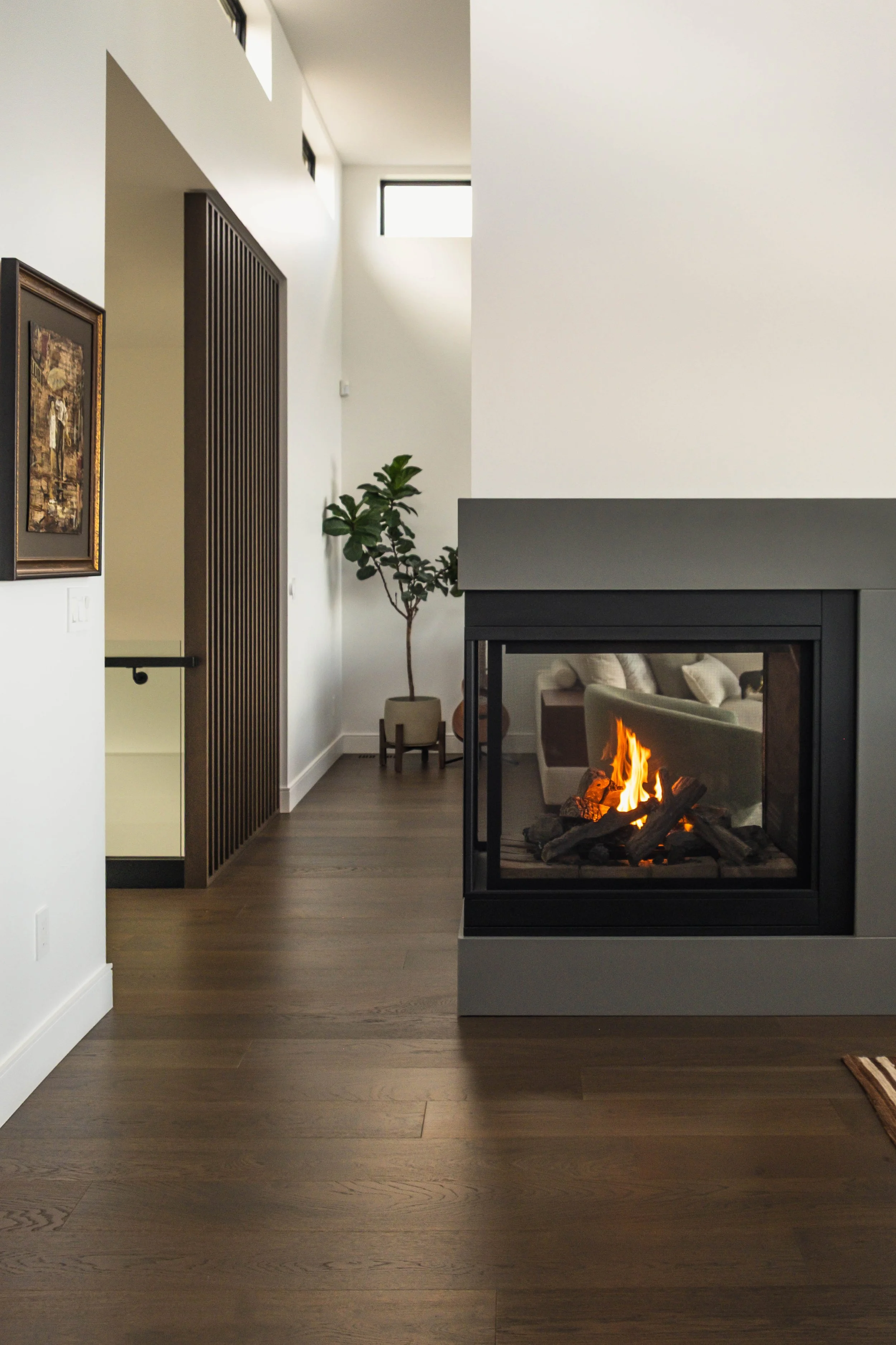 Modern living room with a wood-burning fireplace, hardwood floors, a potted plant, artwork on the wall, and natural light coming through high windows.