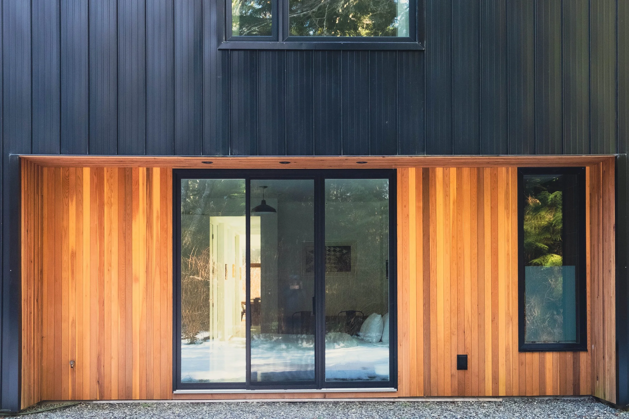 Modern house with black metal siding and a wooden-paneled sliding glass door surrounded by vertical wood planks, with a window above.