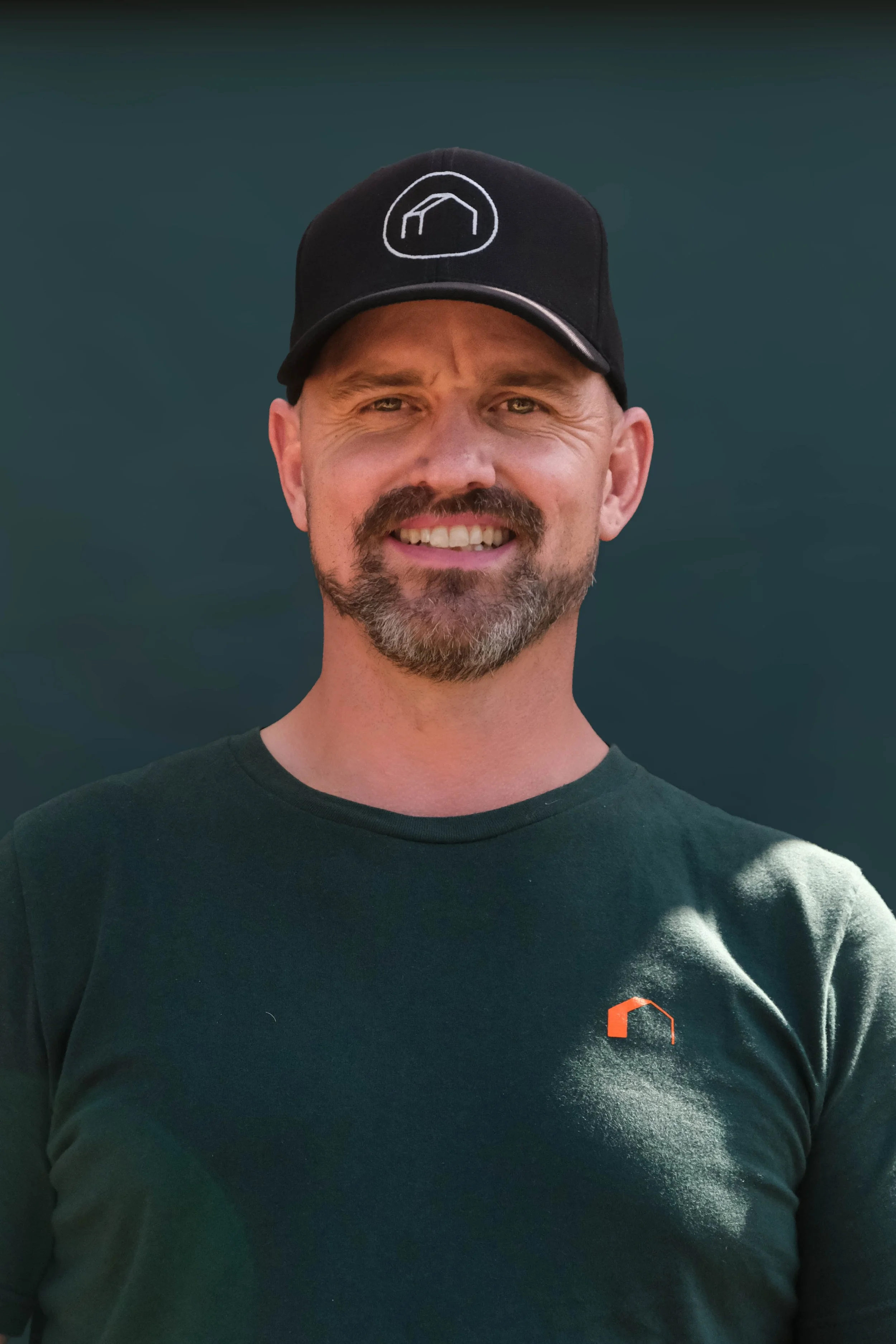 A man with a beard and mustache wearing a black cap with a white logo and a black T-shirt with a small orange logo on the chest, smiling at the camera against a dark background.