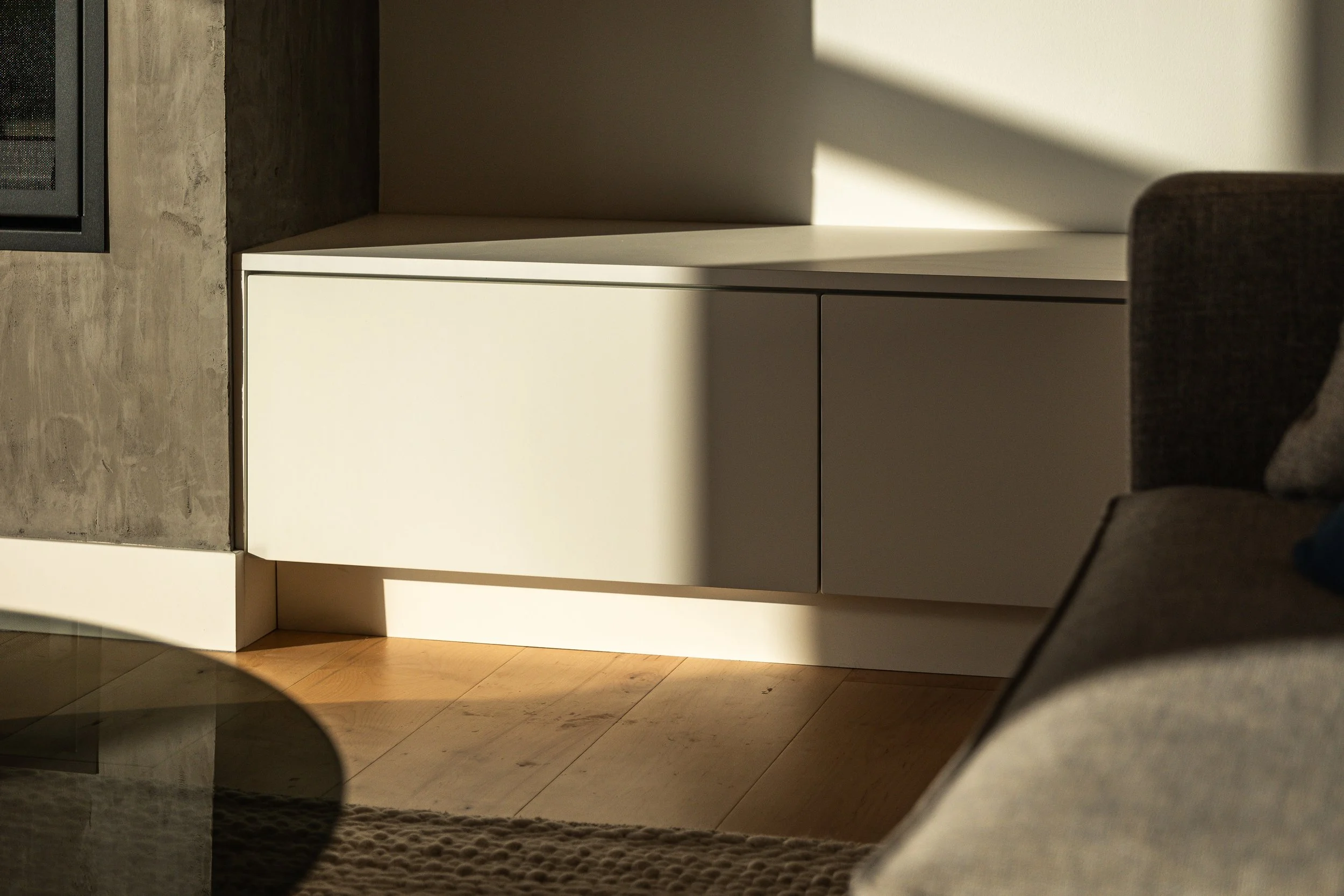 Sunlight casting shadow on a modern white cabinet against a wall with a wood floor and a grey couch to the side.