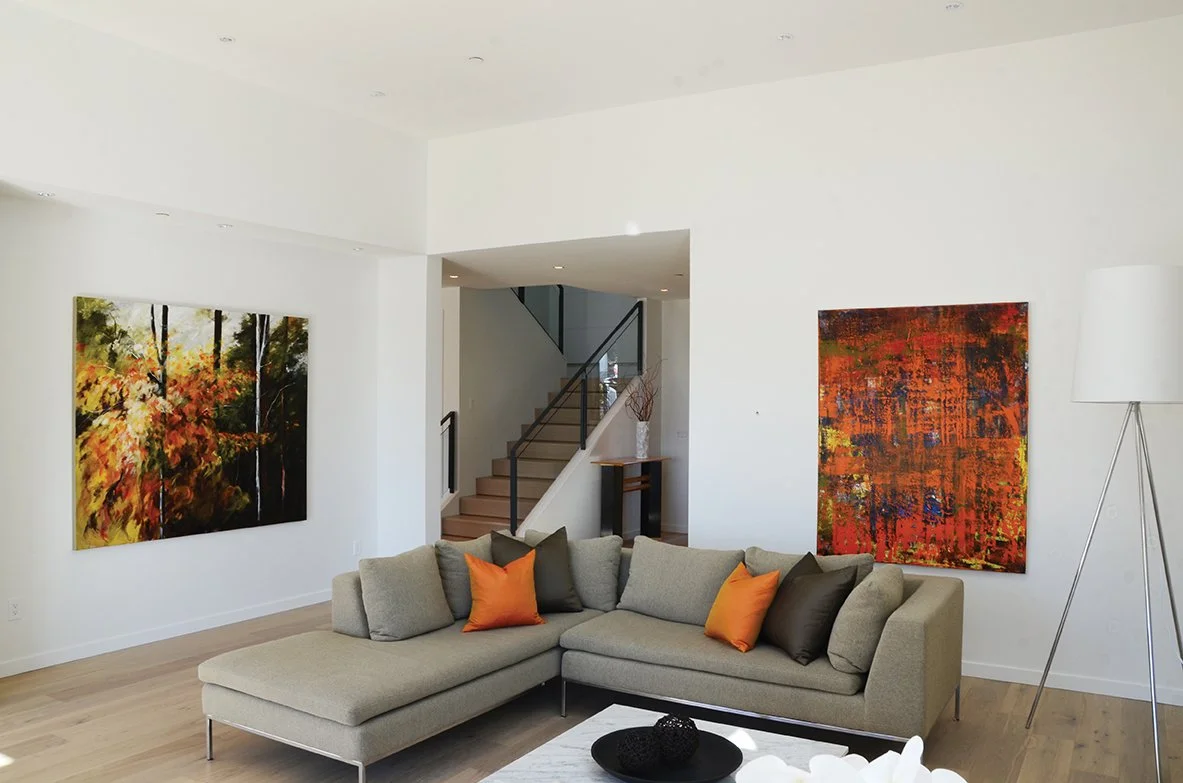 Living room with a beige sectional sofa, orange and dark gray pillows, abstract fall-themed paintings on white walls, light wood flooring, and a white floor lamp.