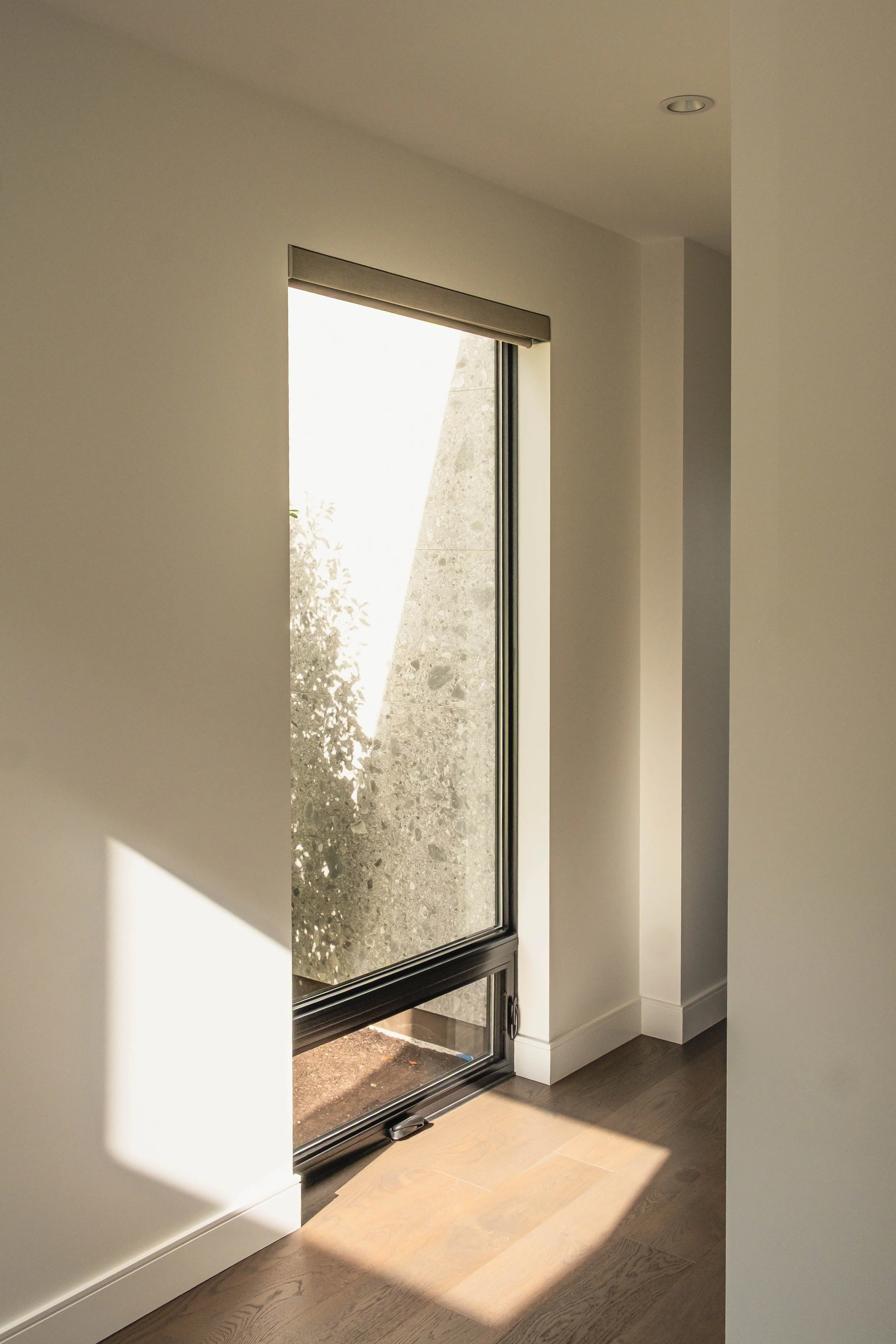 Sunlight entering through a large vertical glass door, casting a shadow on the wooden floor inside a house.